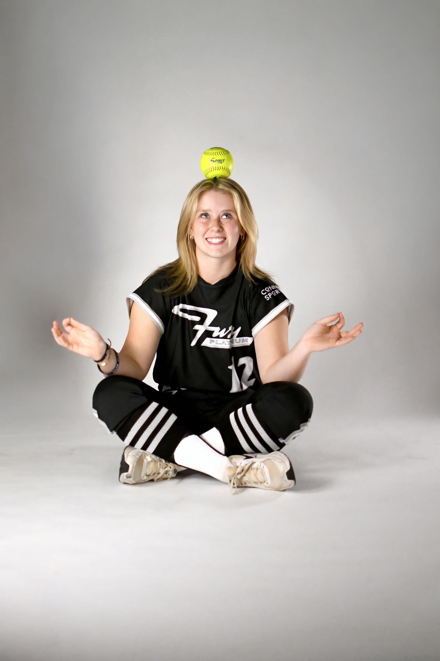 Blonde softball player in black Fury uniform sitting cross-legged with softball balanced on head.