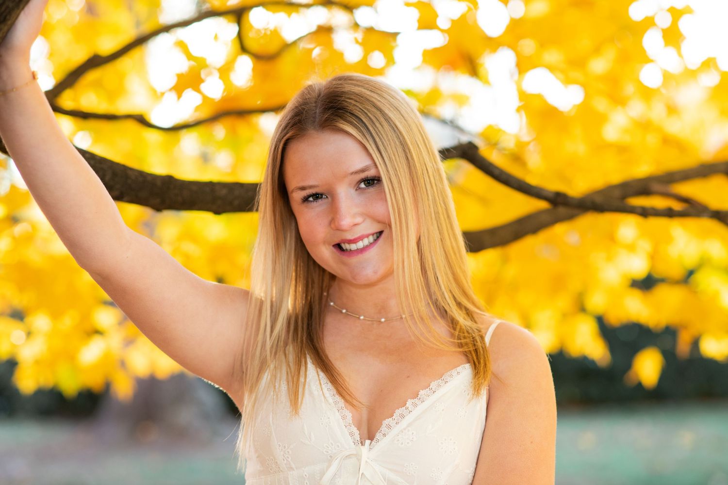Smiling blonde girl in white dress reaching up to golden autumn tree branch for senior portrait.