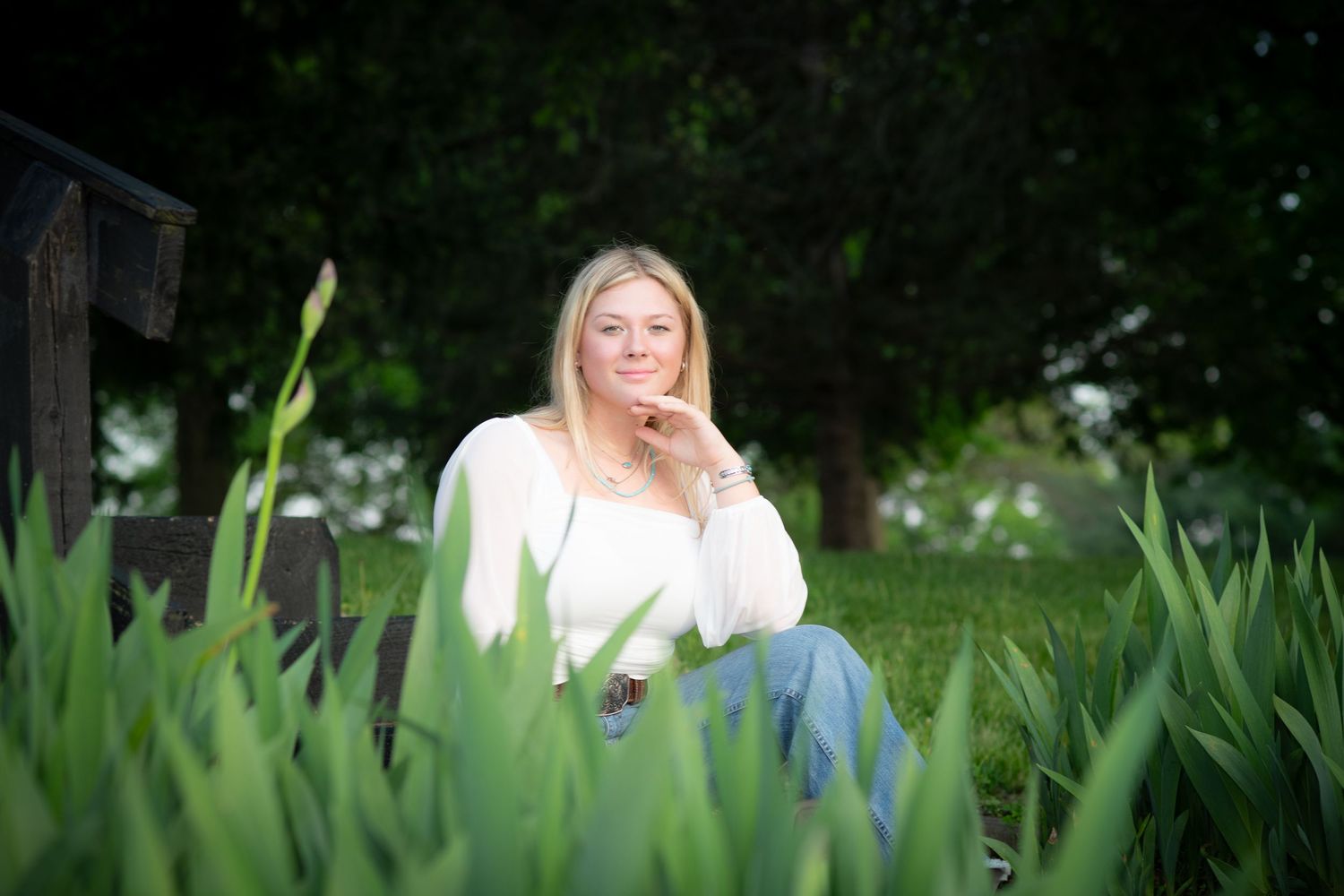 Blonde woman in white top relaxes on bench surrounded by lush green iris plants outdoors.