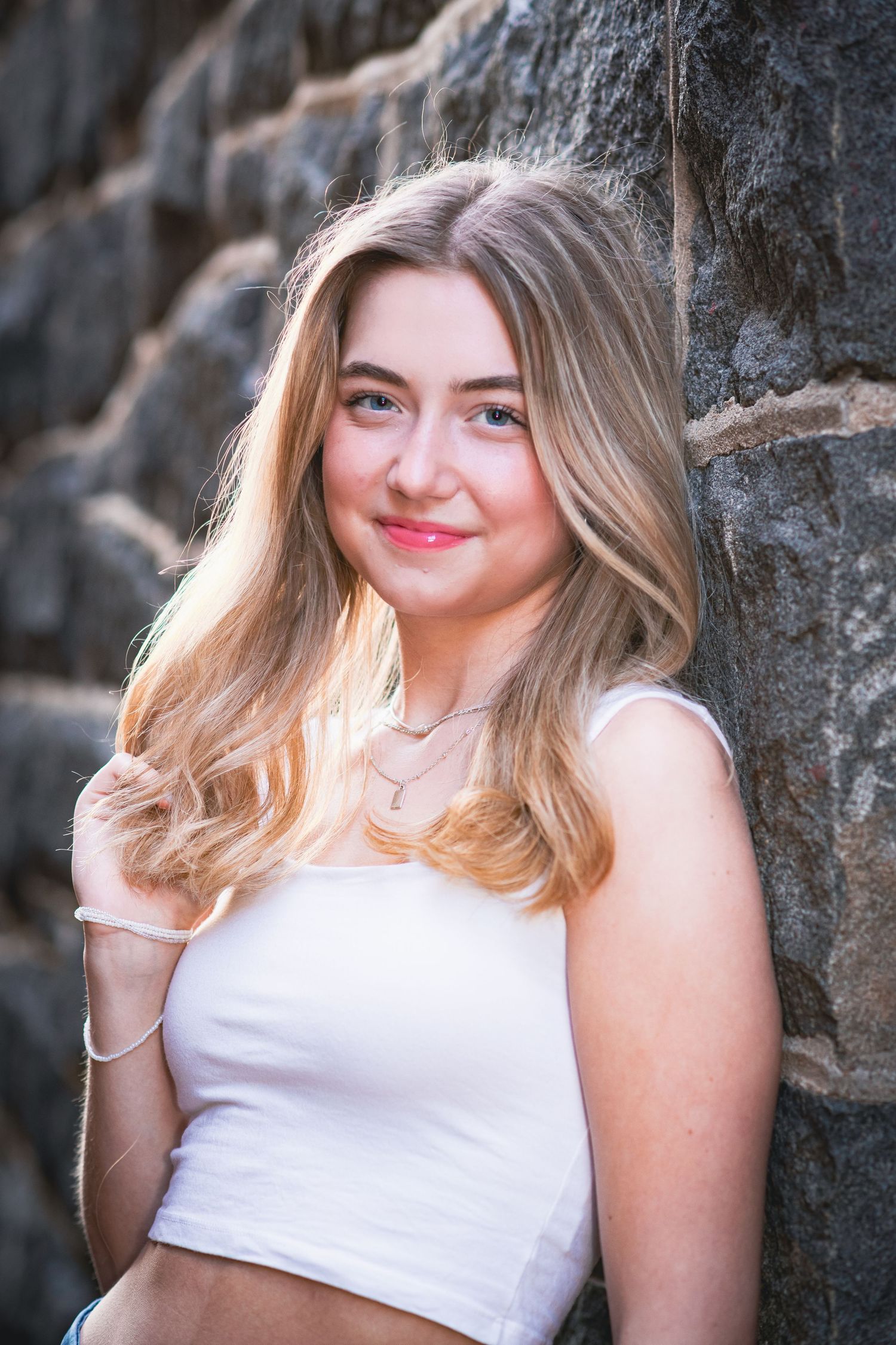 Smiling blonde teenage girl leaning against stone wall wearing white crop top for senior portrait.