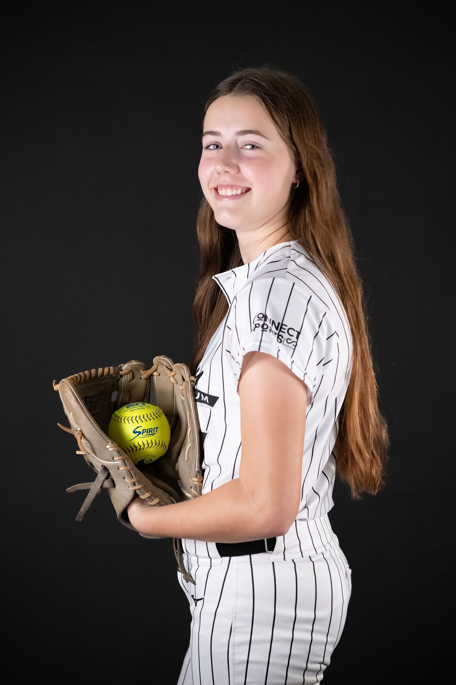 Smiling softball player in pinstripe uniform holding yellow softball in glove against dark background.