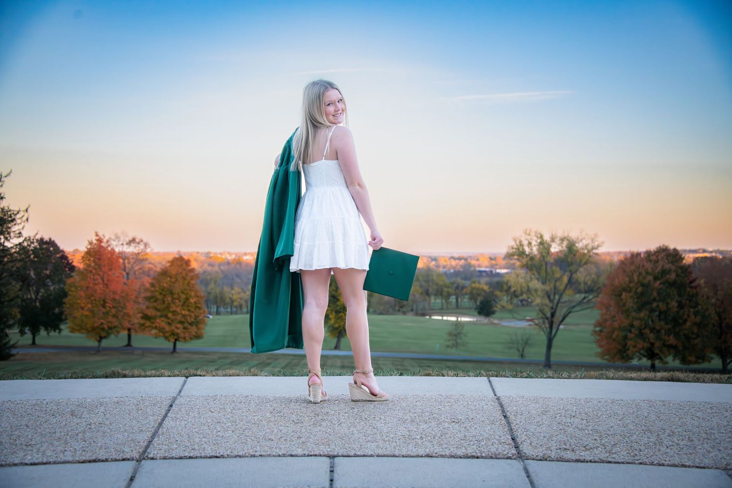Young woman in white dress and green graduation gown poses outdoors at sunset with autumn trees in background.