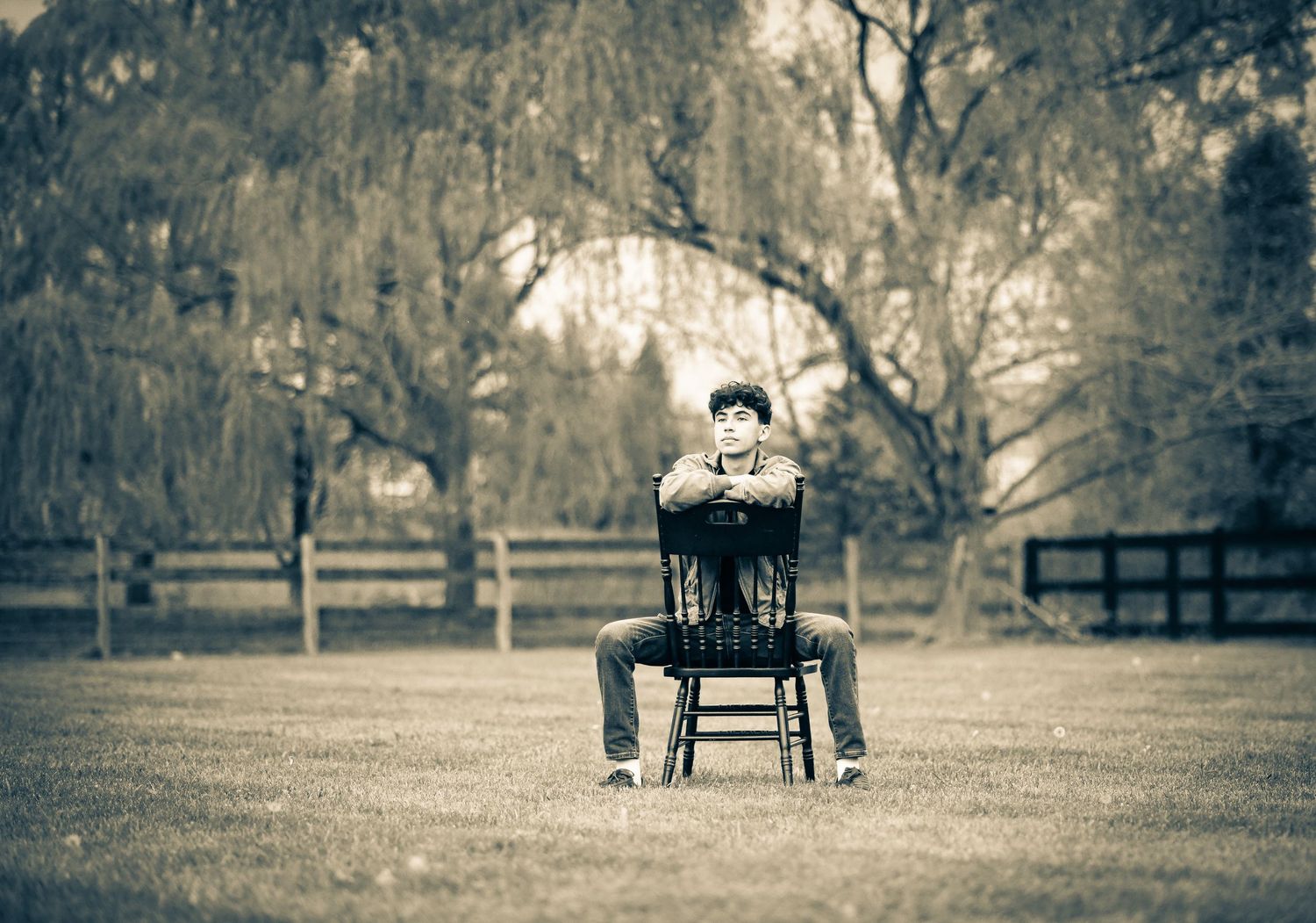 Young man sitting cross-armed on a wooden chair in an open field with weeping trees, sepia-toned portrait.