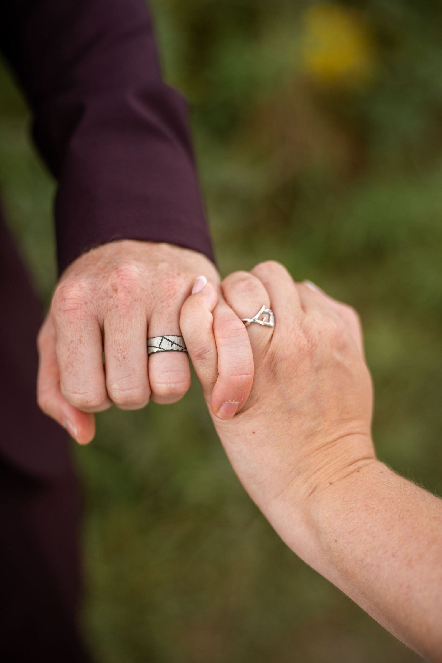 Close-up of newlyweds holding hands displaying matching silver wedding rings against green background.