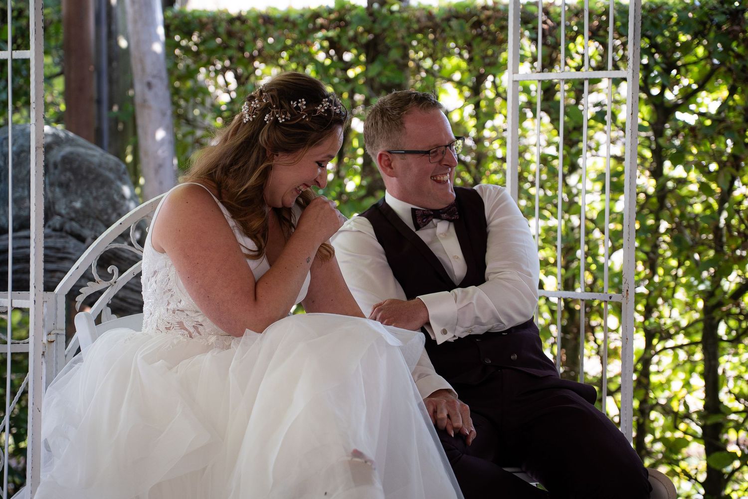 Bride laughs joyfully while groom smiles beside her on bench during intimate outdoor wedding moment.