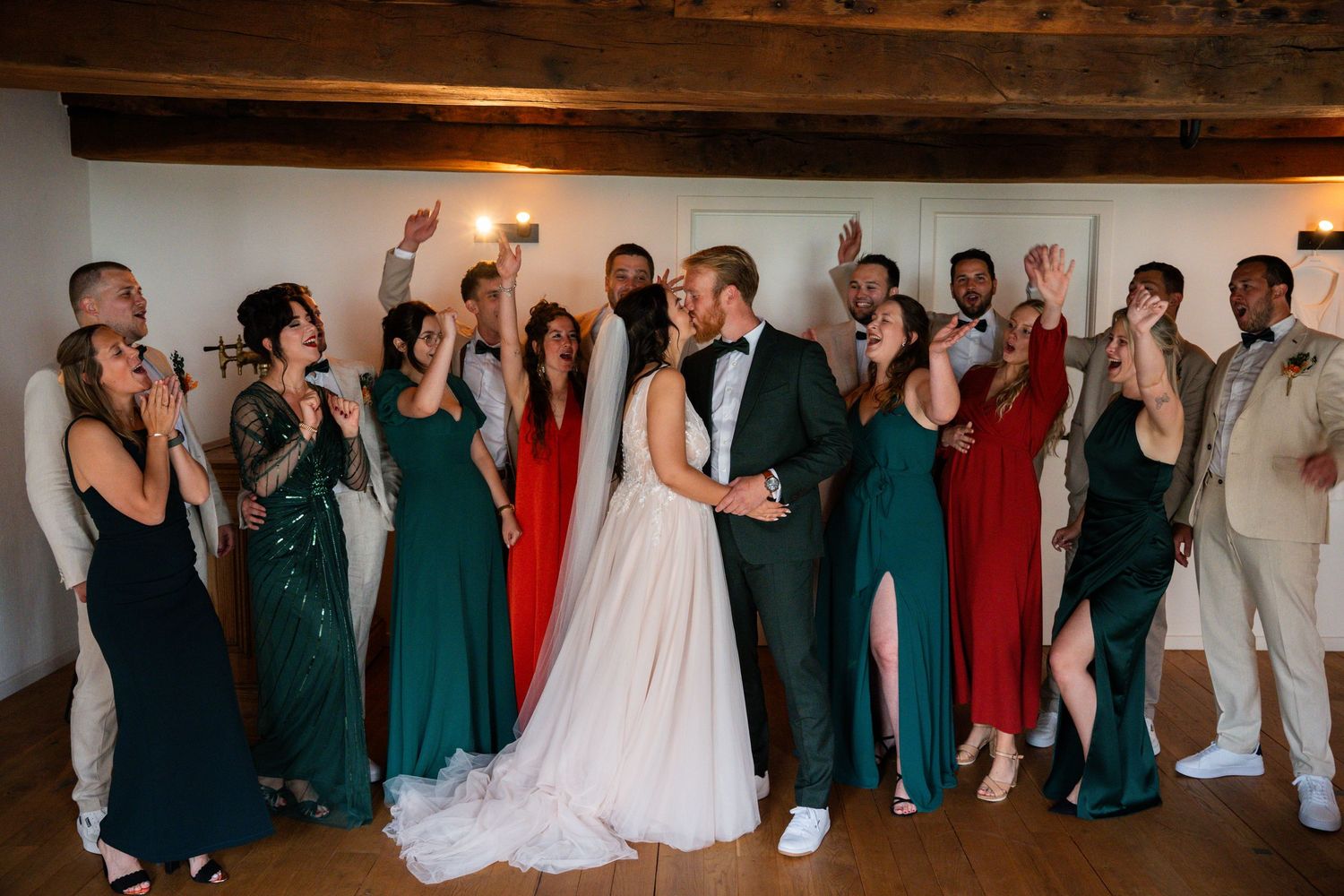 Bride and groom kissing surrounded by cheering wedding party in green and red dresses.