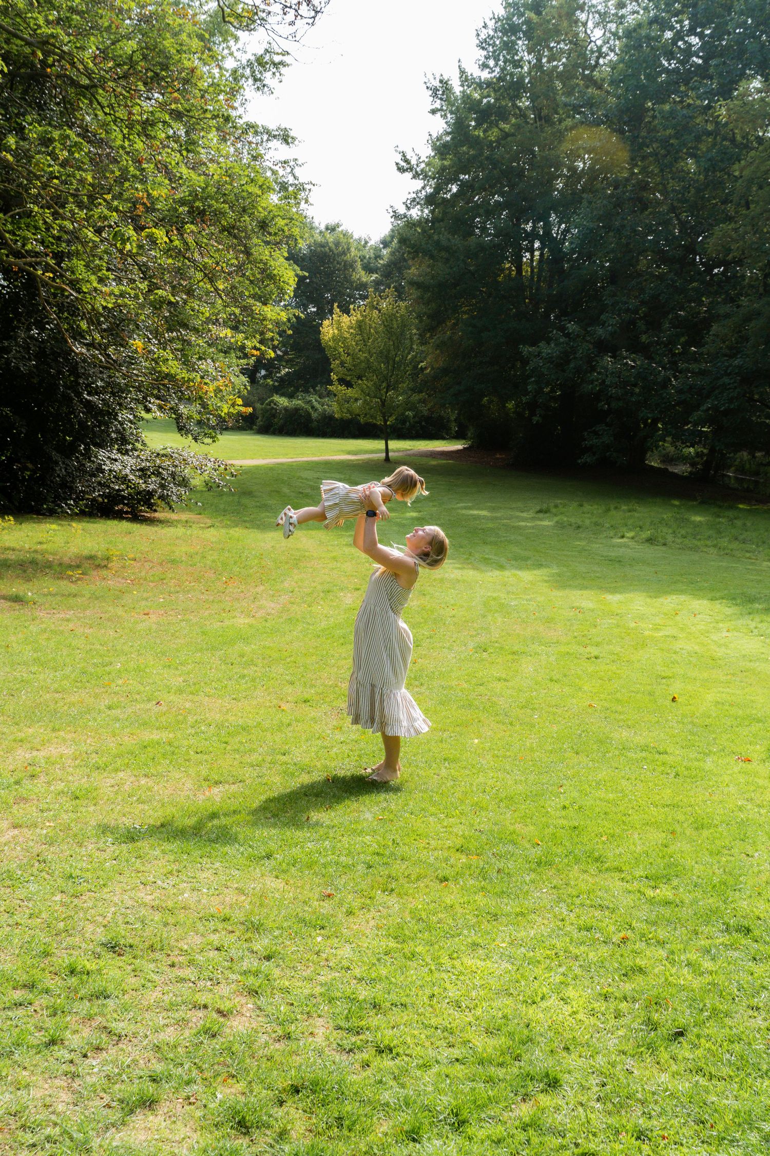 Woman in white dress lifts smiling toddler high in the air in a bright sunlit green park.