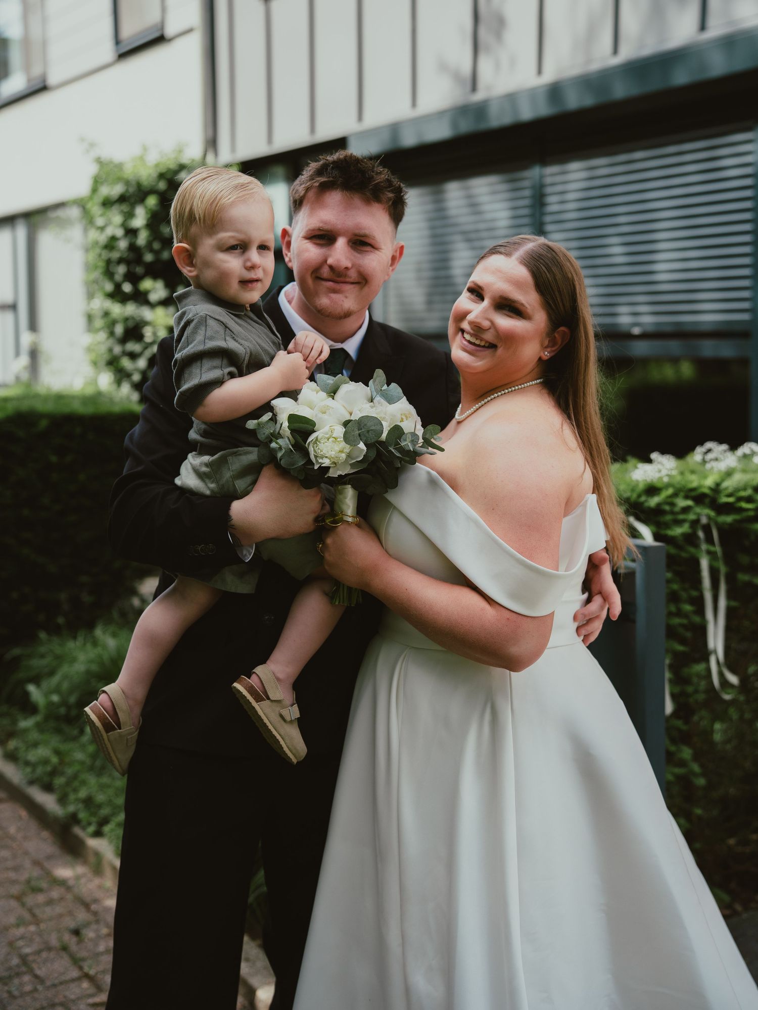 Bride in off-shoulder white gown and groom in black suit holding toddler boy at wedding.
