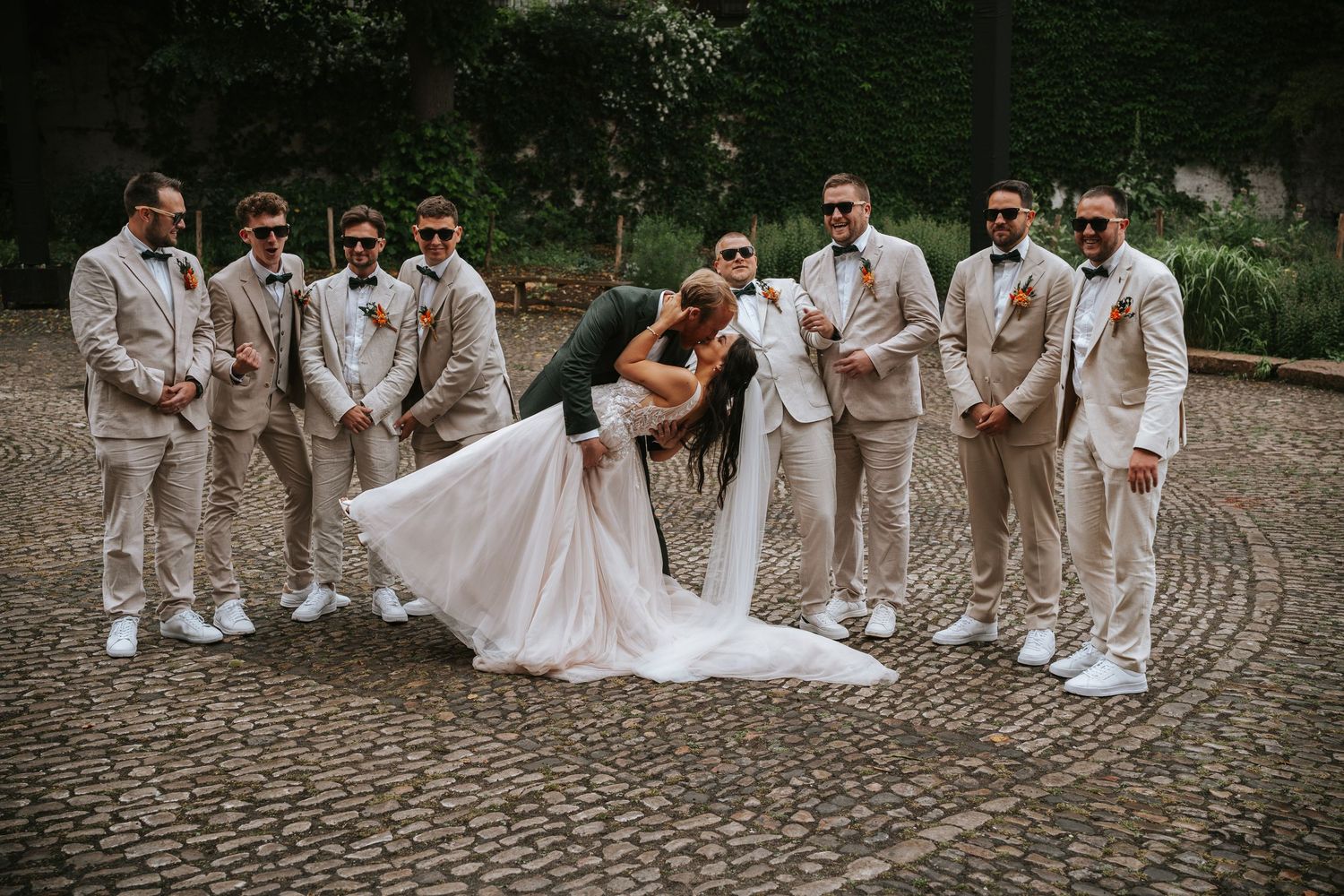 Groom dipping bride in a romantic pose surrounded by groomsmen on a cobblestone plaza.