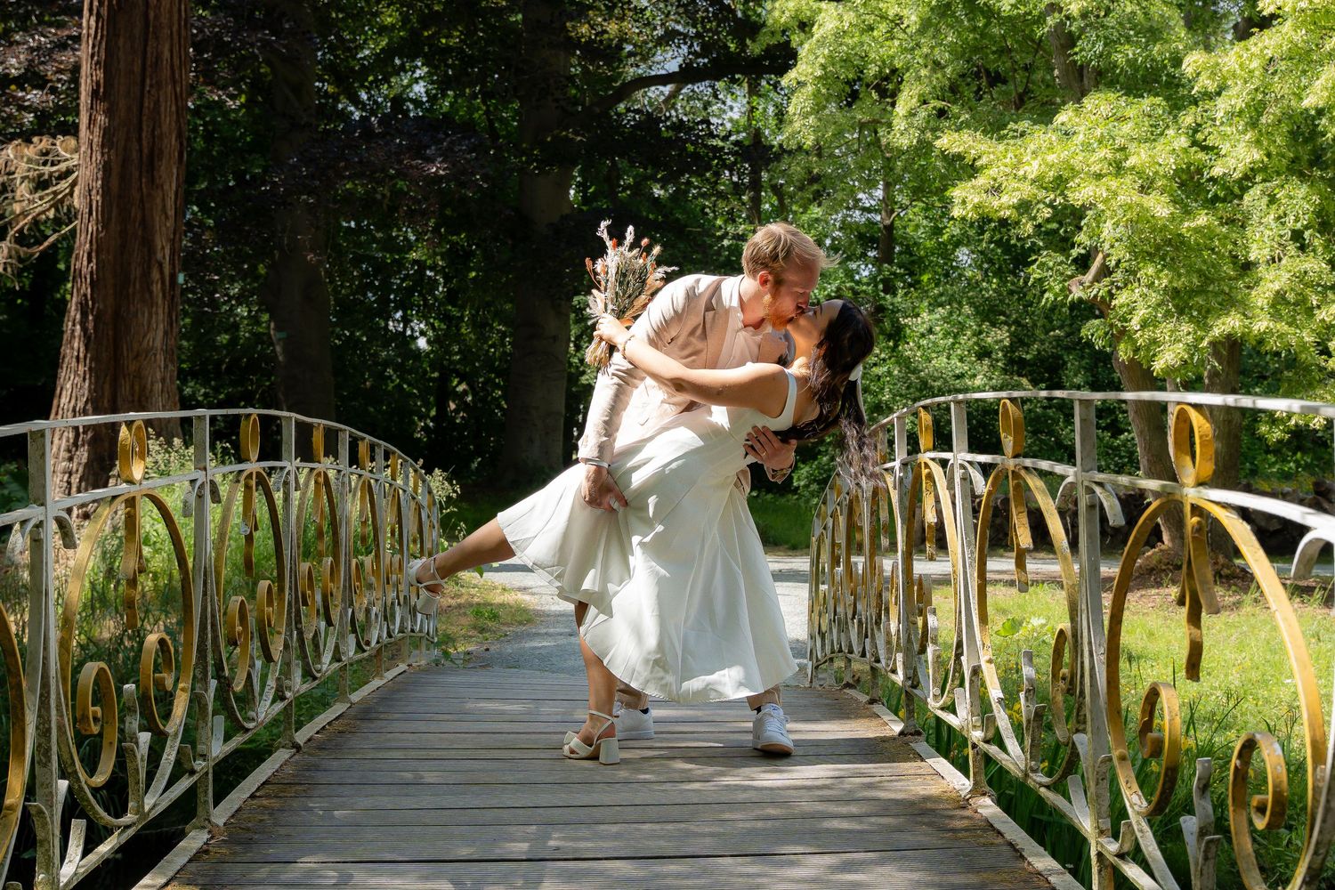 Couple sharing a romantic dip kiss on an ornate bridge surrounded by lush green trees.
