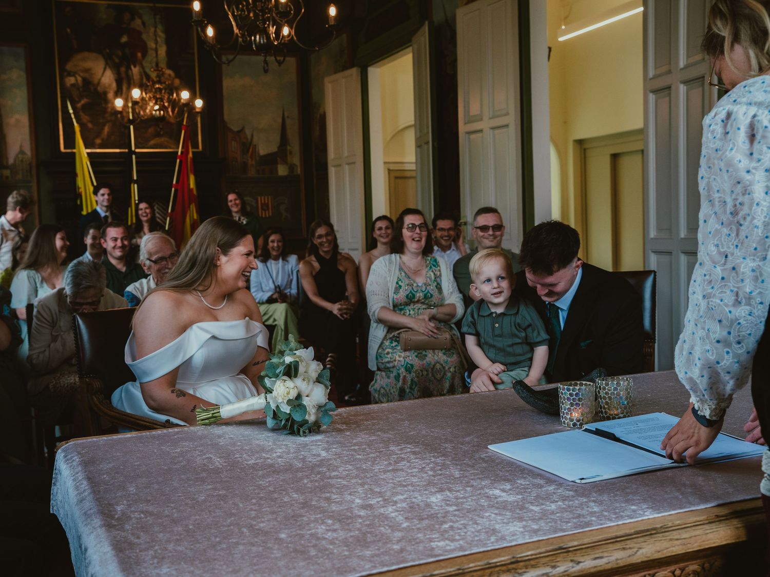 Bride and groom signing wedding register with child at ornate ceremony hall.