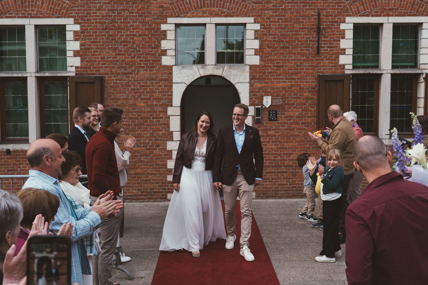 Bride and groom walk down red carpet aisle outside brick building surrounded by cheering wedding guests.