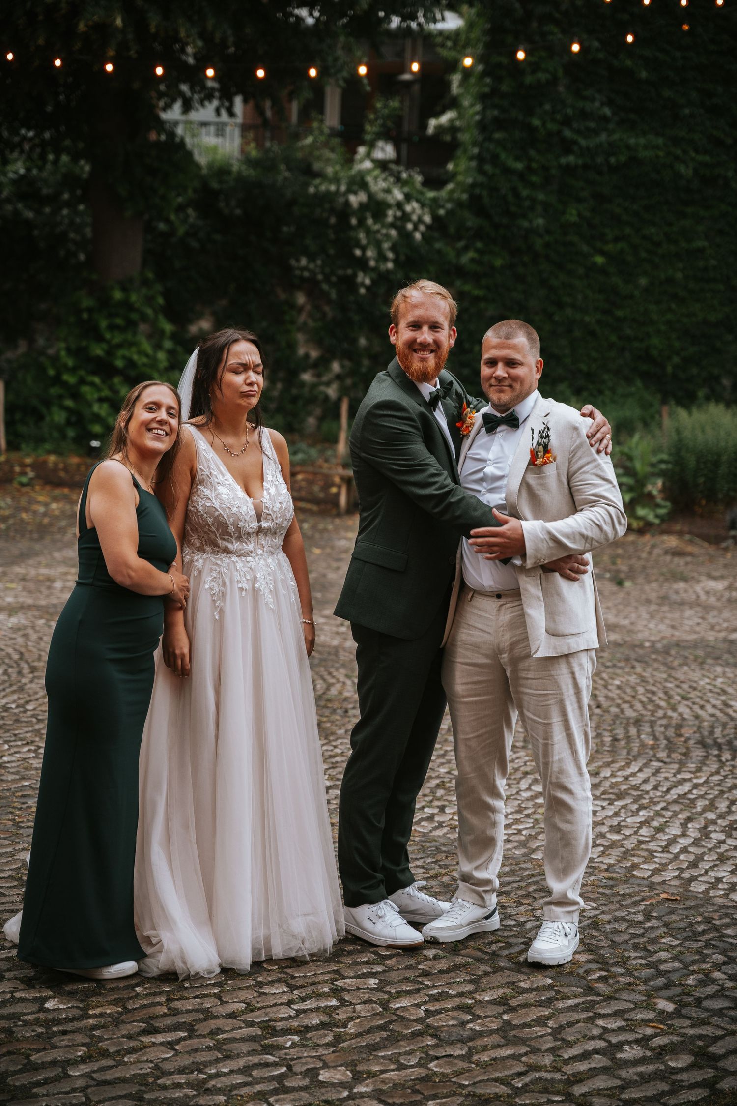 Wedding party of four posing on cobblestone courtyard with string lights and ivy backdrop.