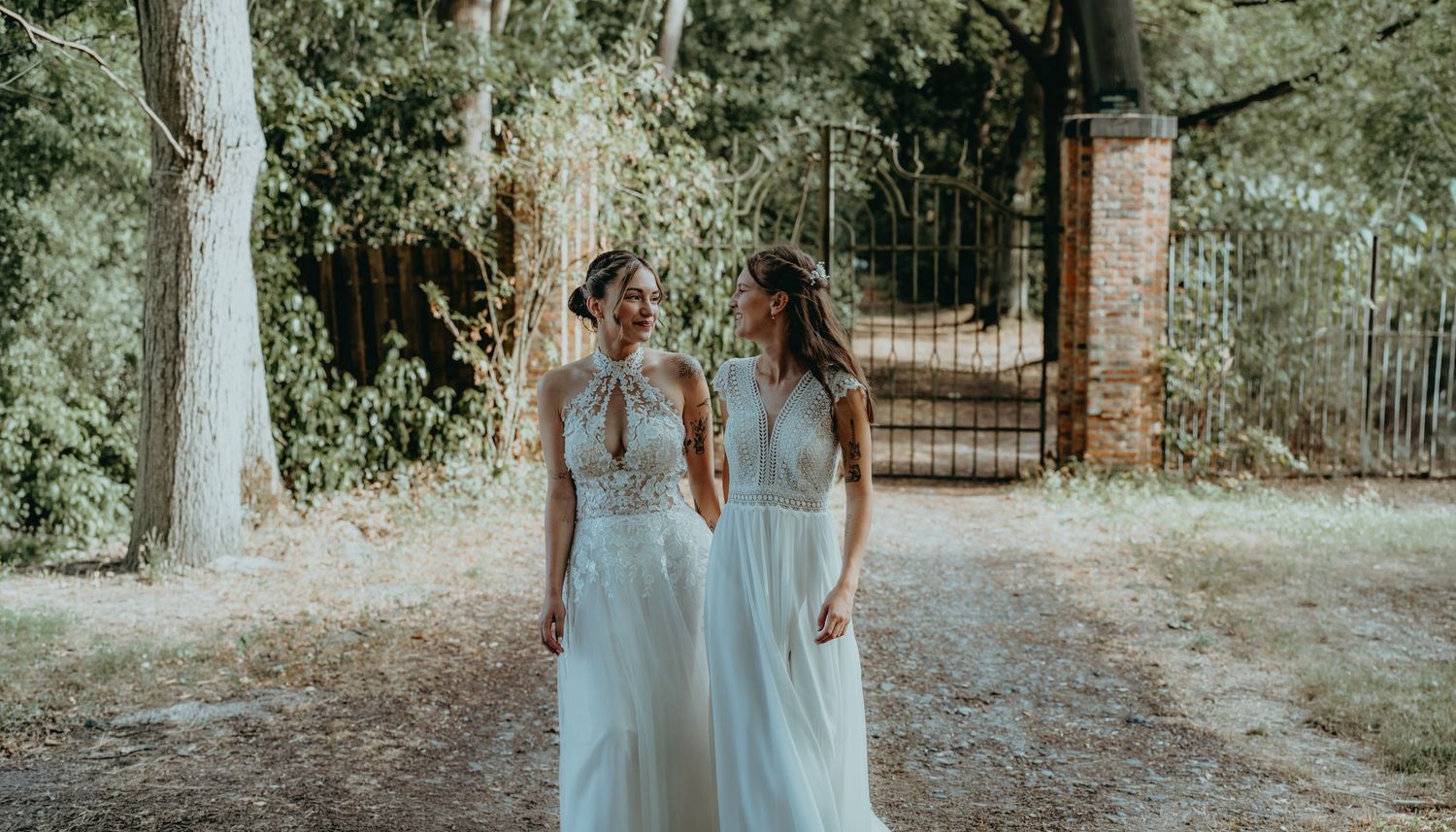 Two brides in flowing white wedding dresses walk together smiling near a brick gate in wooded setting.