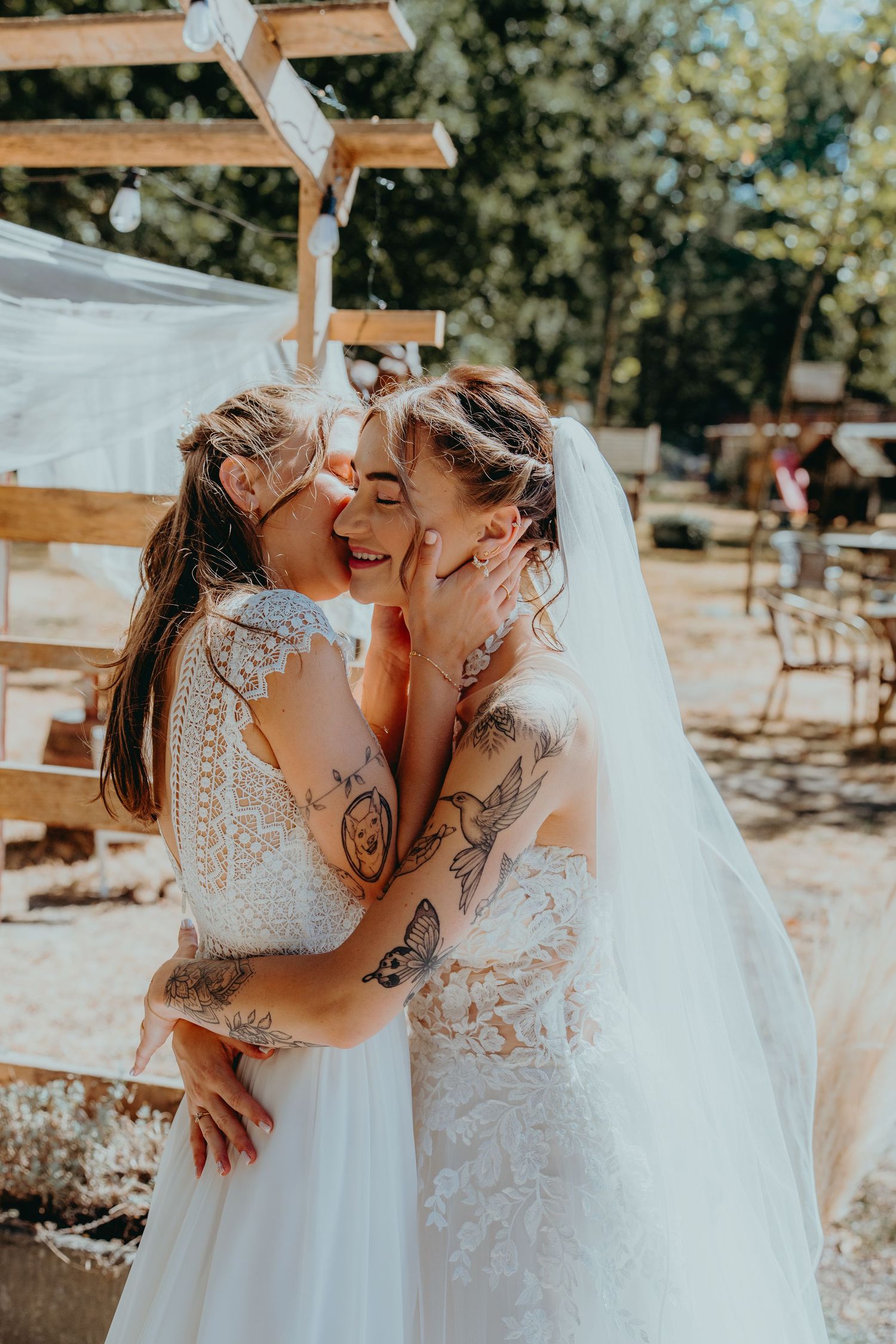 Two tattooed brides in white wedding dresses sharing a tender kiss at outdoor boho wedding ceremony.