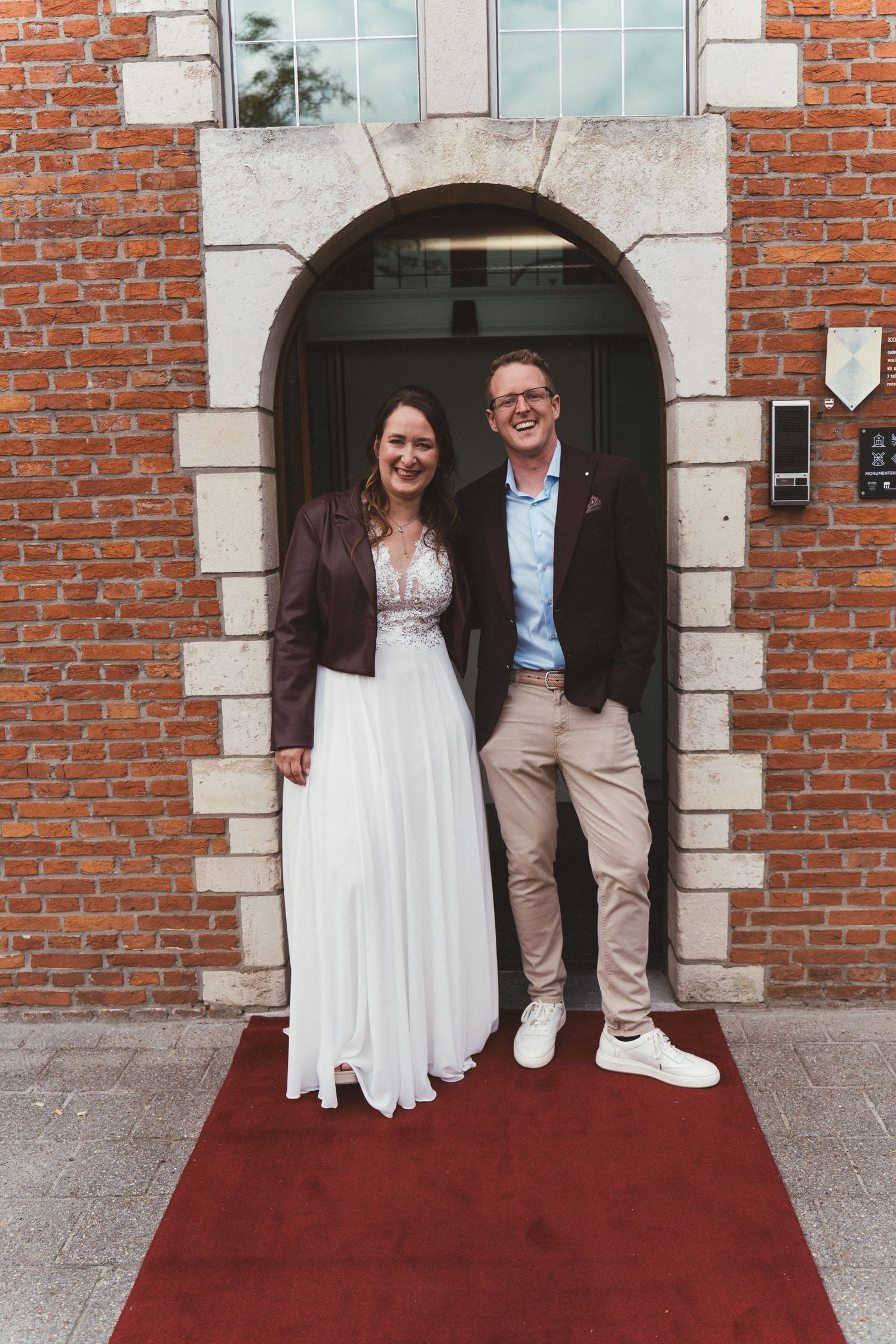 Smiling couple in wedding attire standing on a red carpet under a brick archway entrance.