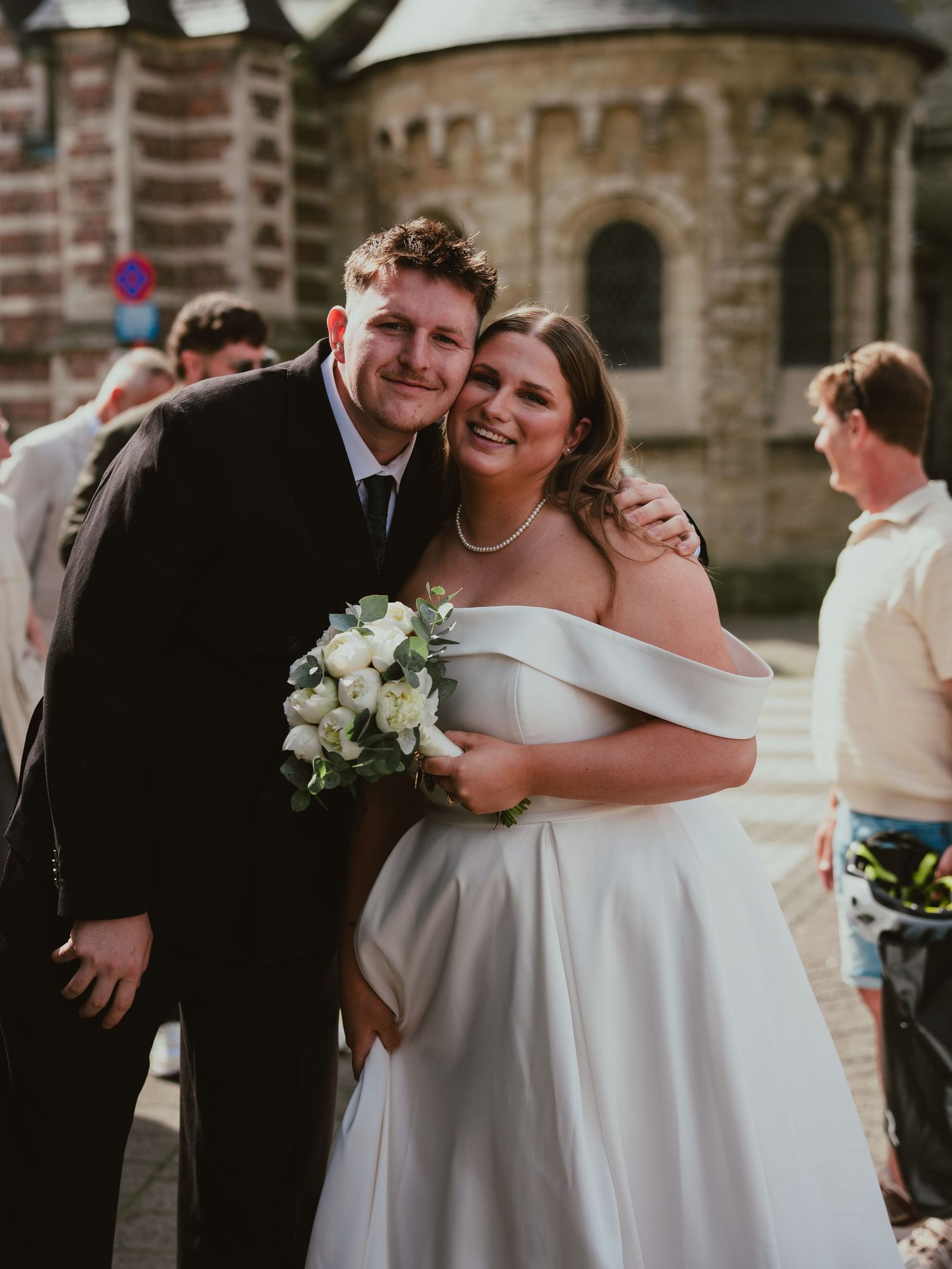 Smiling newlyweds embrace outside church, bride holding white floral bouquet in elegant gown.