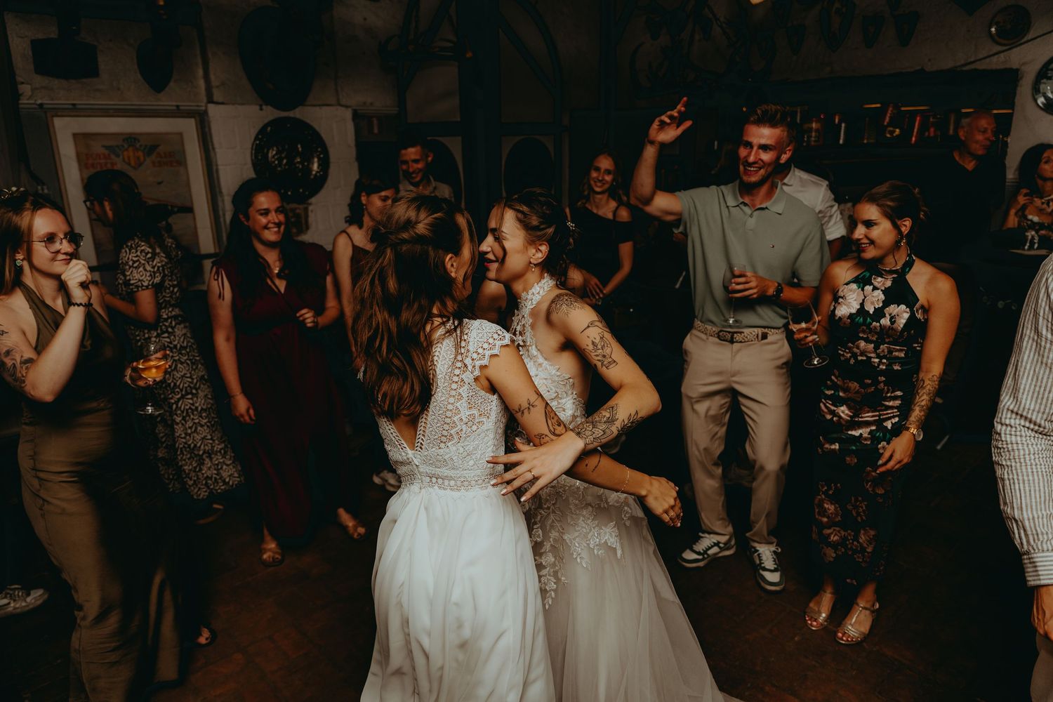 Two brides in white wedding dresses dancing joyfully with guests at their lively wedding reception.
