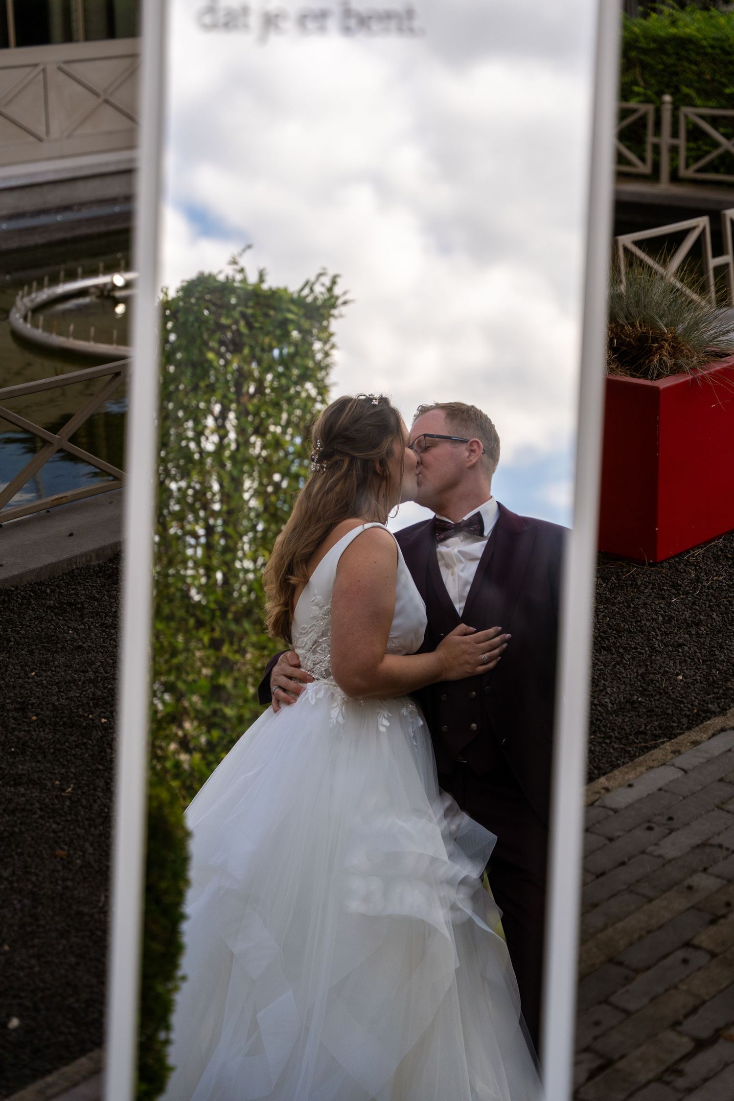 Wedding couple kissing reflected in tall mirror with Dutch text at outdoor venue on cloudy day.