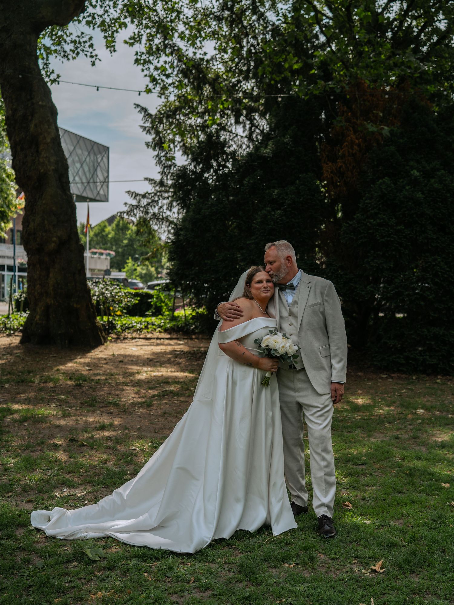 Bride in flowing white gown and groom in tan suit pose romantically in garden.