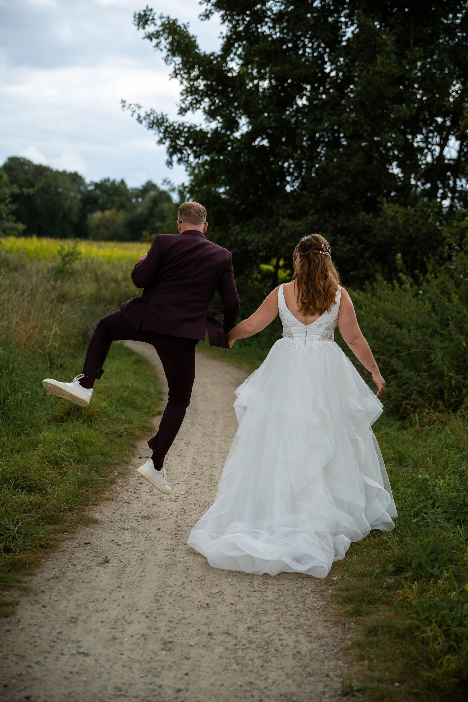 Joyful groom jumps while holding bride's hand as couple walks down rural path in wedding attire.
