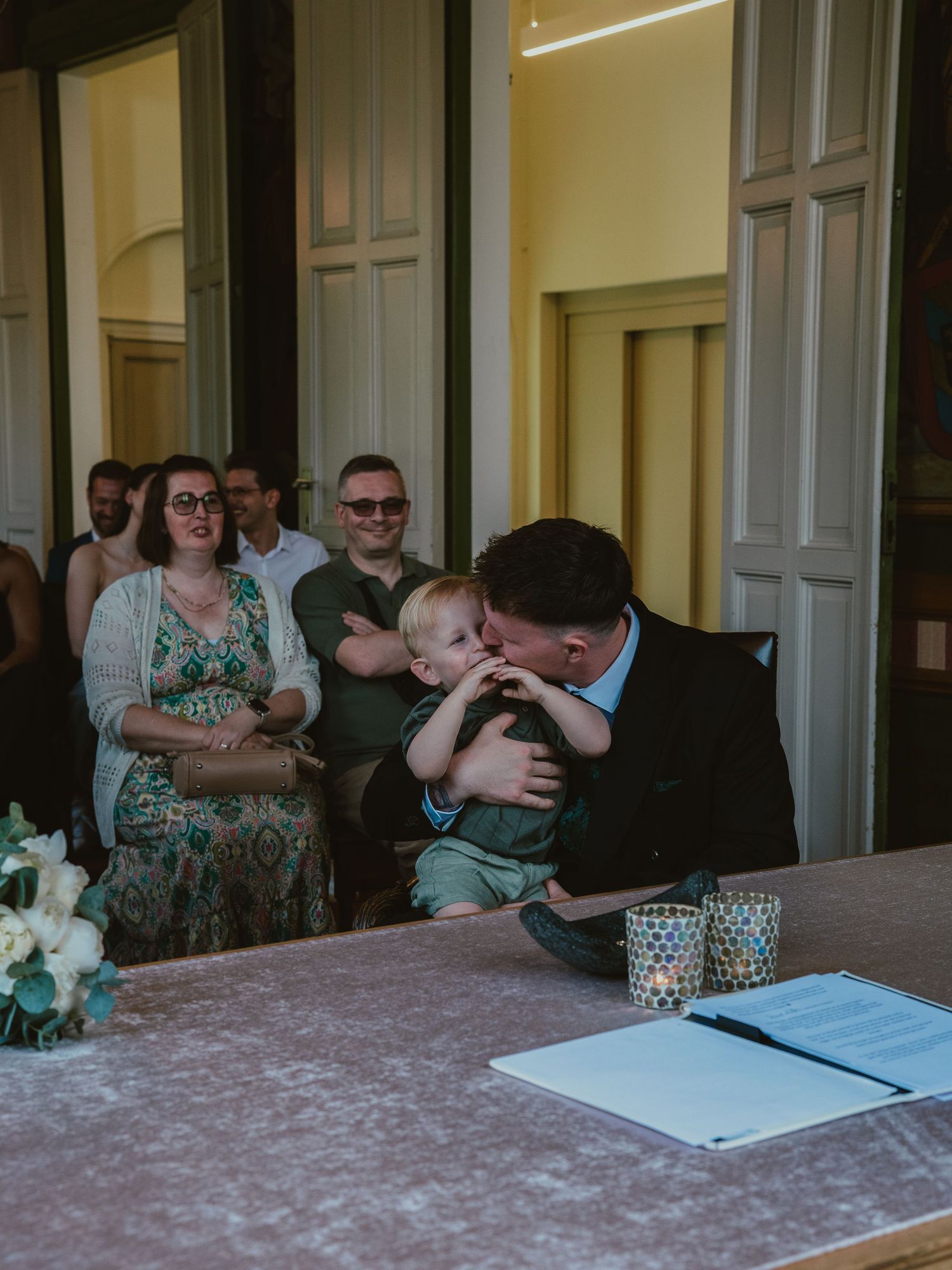 Groom in black suit shares tender moment kissing young child at wedding ceremony.