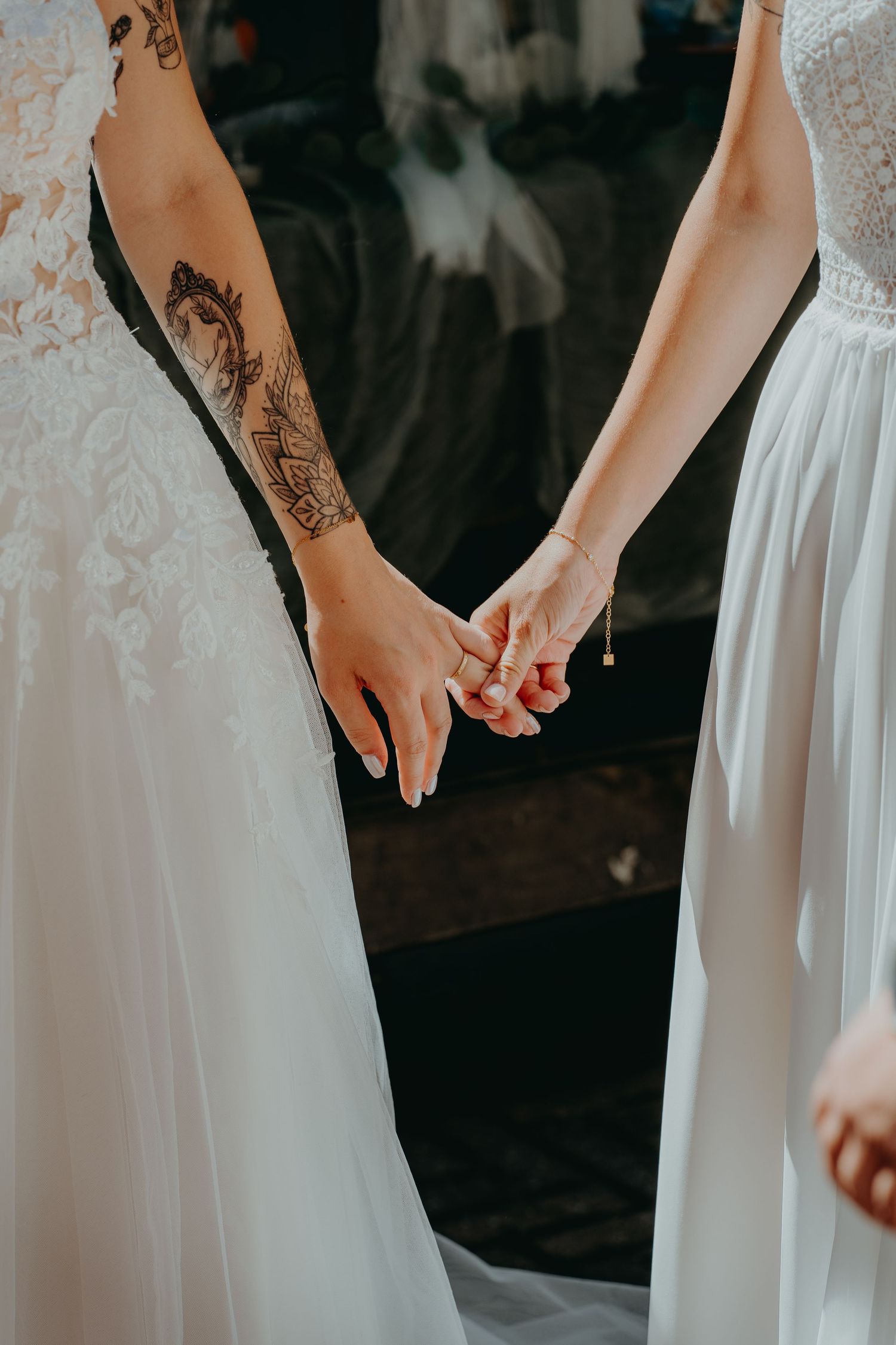 Two brides in white wedding dresses hold hands, one arm adorned with an elaborate tattoo sleeve.