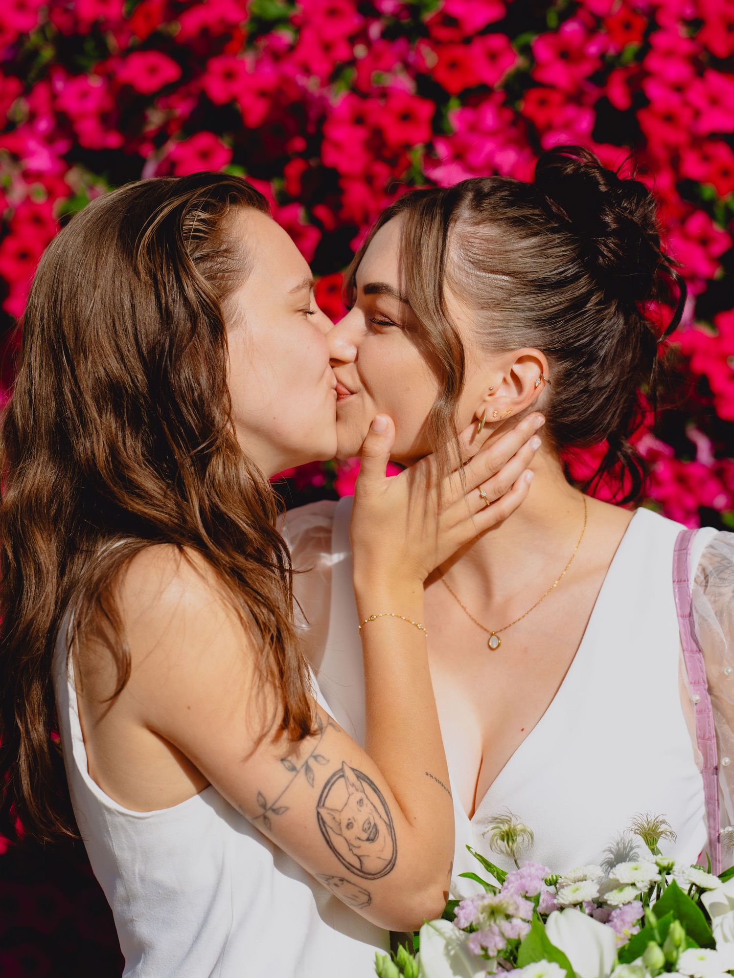Two brides sharing a romantic kiss in front of vibrant pink flowers on their wedding day.