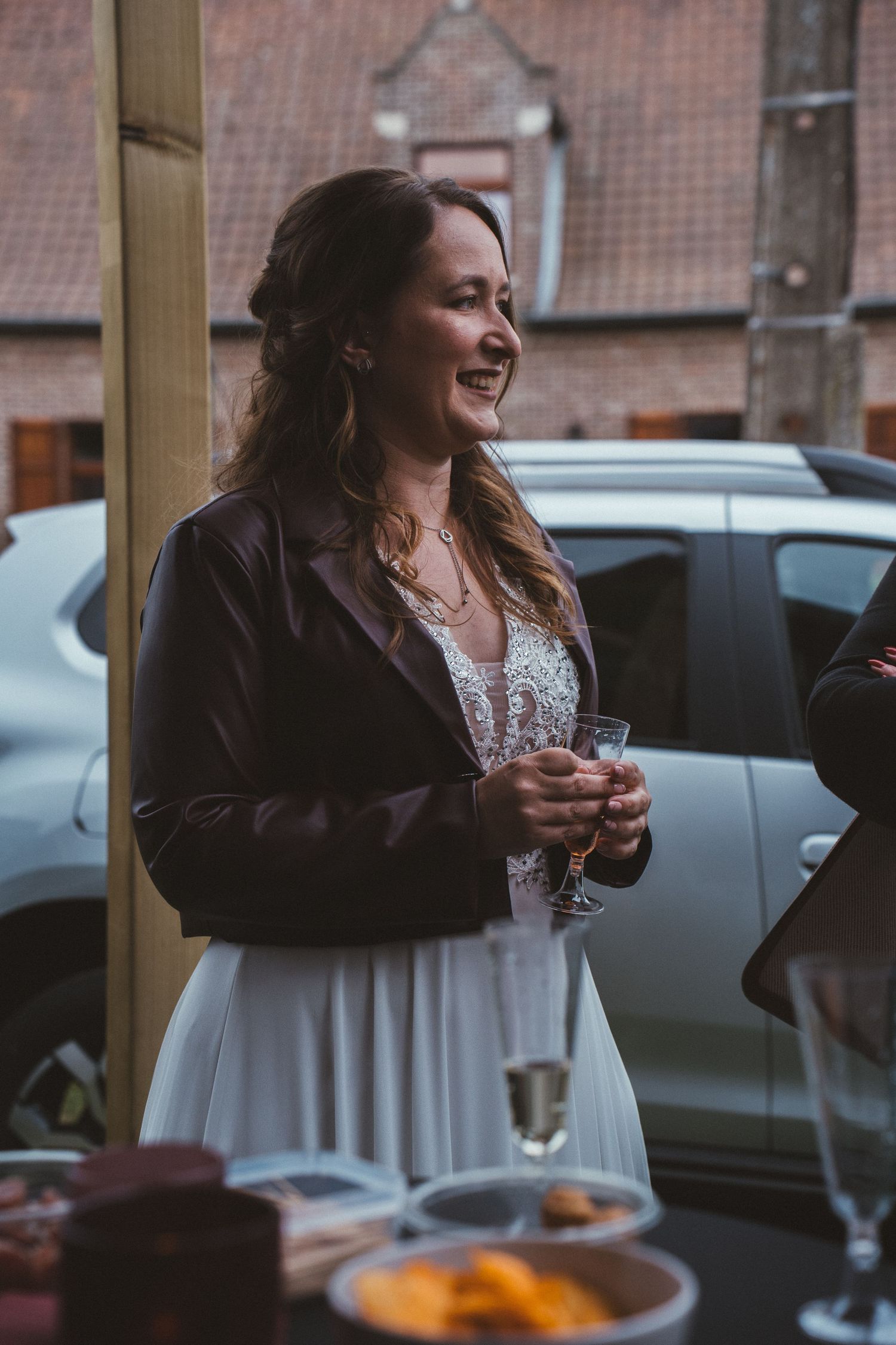 Smiling bride in white dress and leather jacket holds champagne glass at outdoor wedding reception.