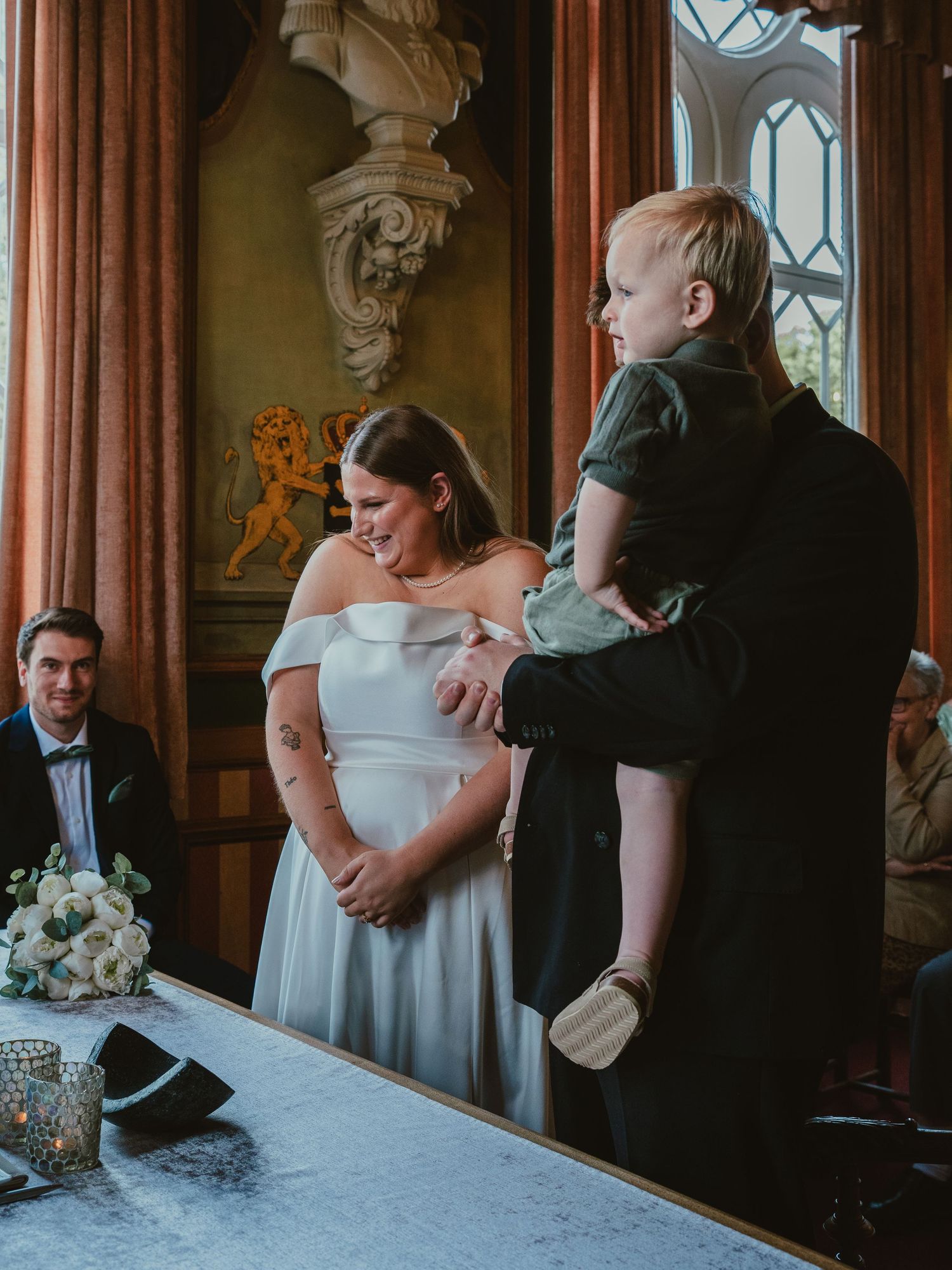 Bride in white off-shoulder gown laughs as groom holds toddler during indoor wedding ceremony.