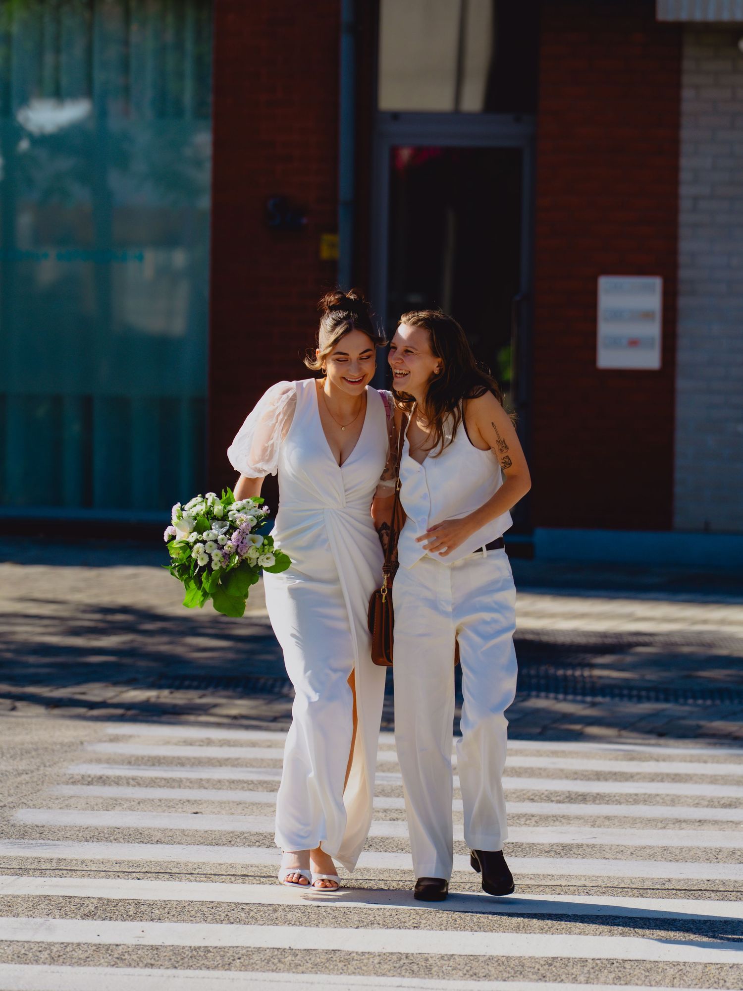 Two women in white outfits walk together across street, one holding floral bouquet.