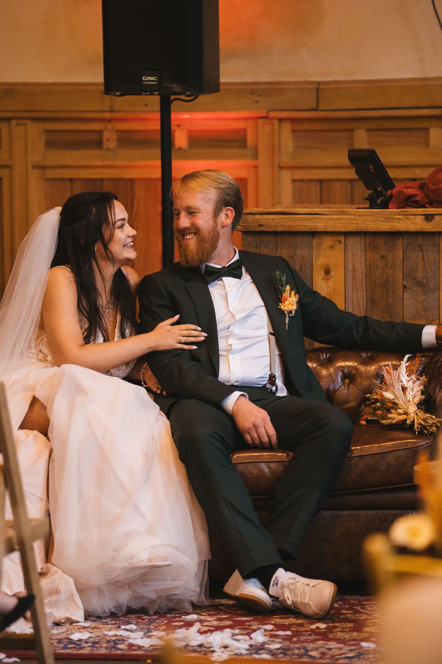 Bride and groom laughing together on a leather sofa during their indoor wedding reception.