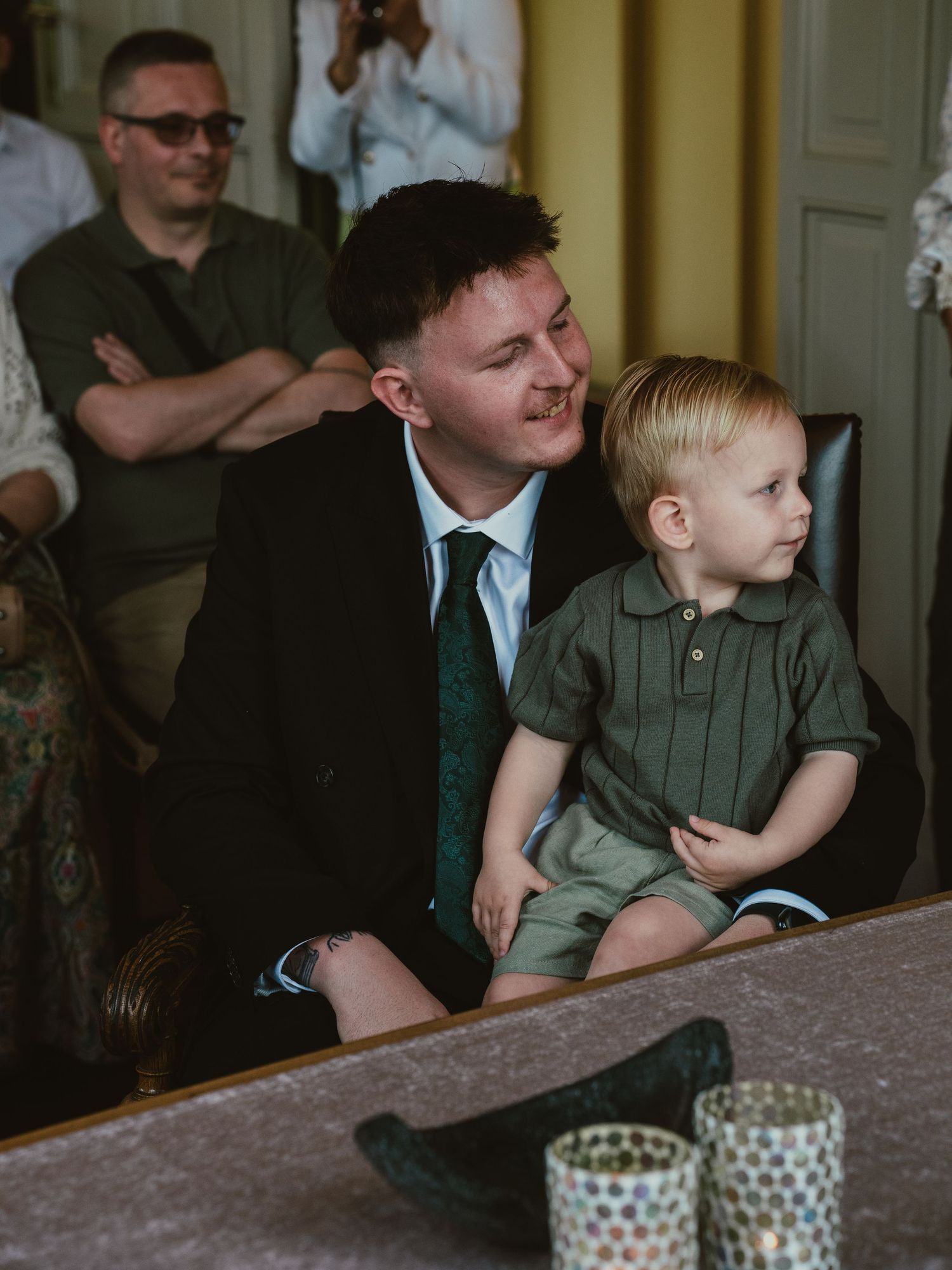 Groom in black suit smiles at young blonde toddler boy during wedding ceremony.