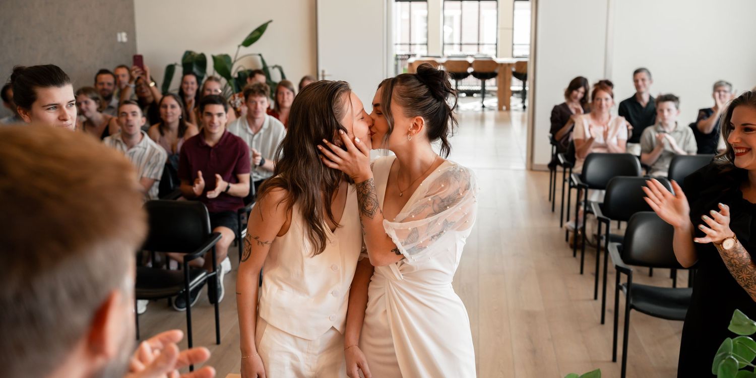 Two brides share a first kiss at their wedding ceremony as guests applaud in a modern venue.