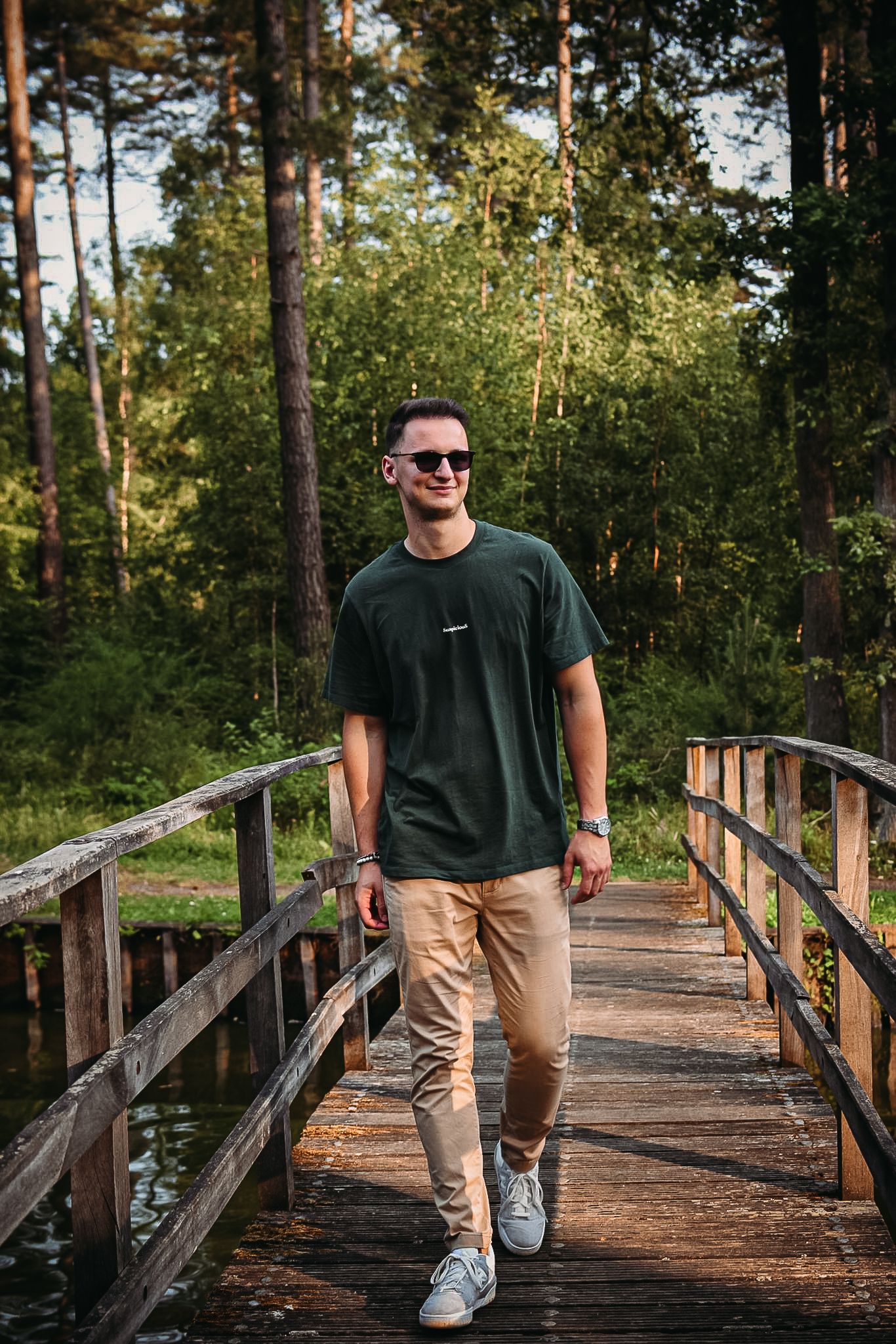 Smiling young man in green t-shirt and sunglasses walking on a wooden bridge in a forest.