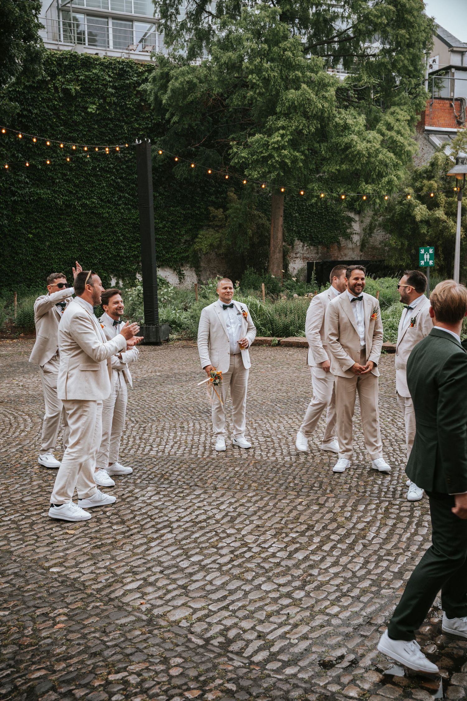 Groomsmen in matching beige suits and white sneakers gathered on a cobblestone courtyard.