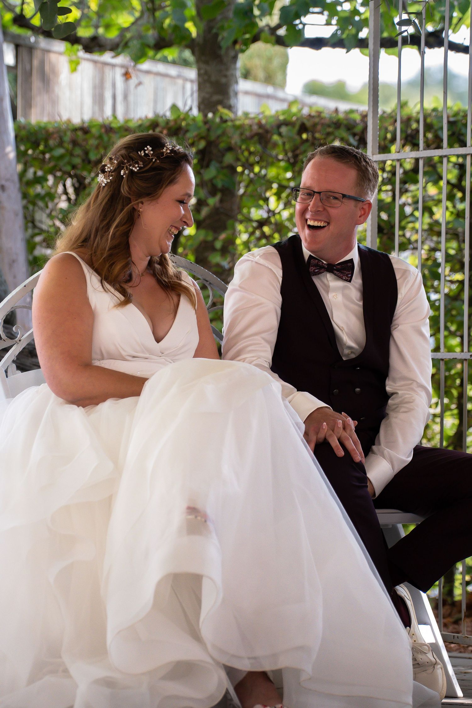Bride and groom laugh together while seated under garden pergola during sunny outdoor wedding celebration.