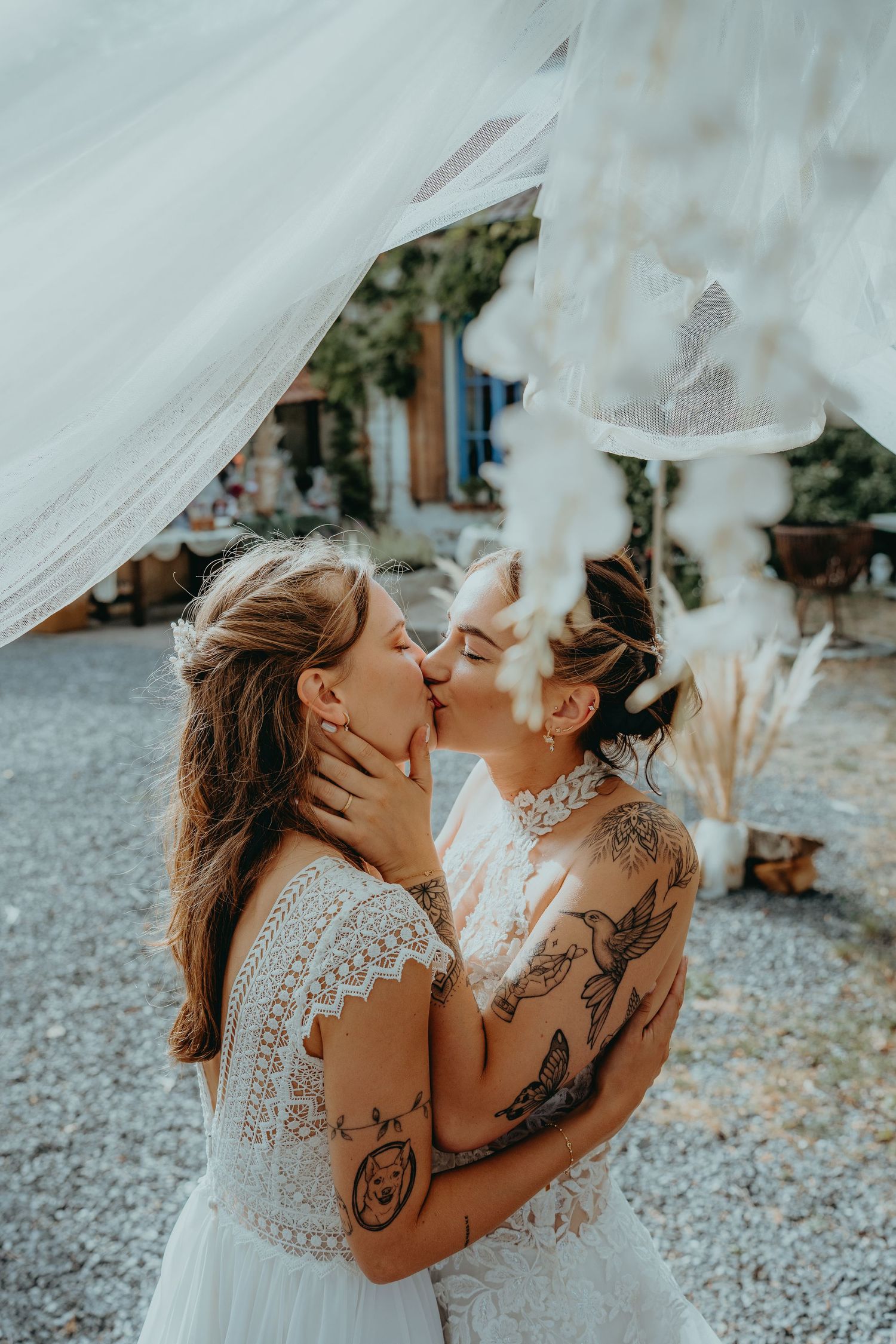 Two tattooed brides in white lace wedding dresses sharing a passionate kiss at boho outdoor wedding.
