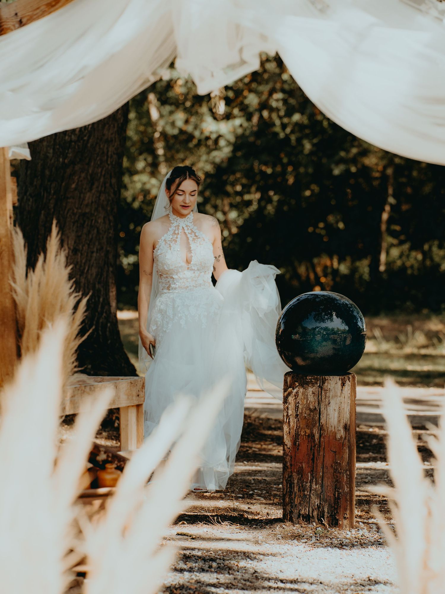 Bride in elegant white lace wedding gown with veil walking through a boho outdoor ceremony arch.
