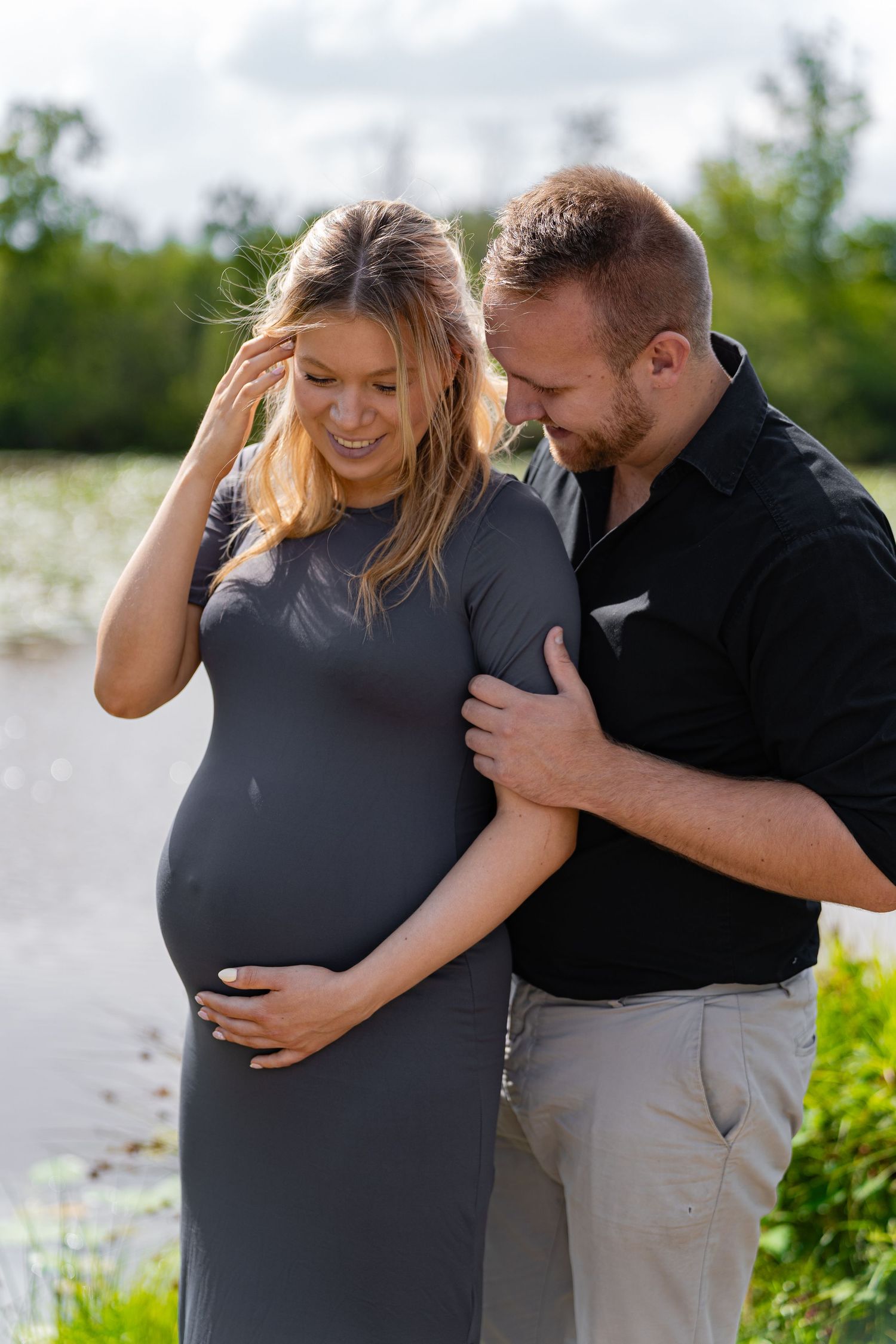 Pregnant couple embraces tenderly outdoors near a lake, man gently touching woman's baby bump.