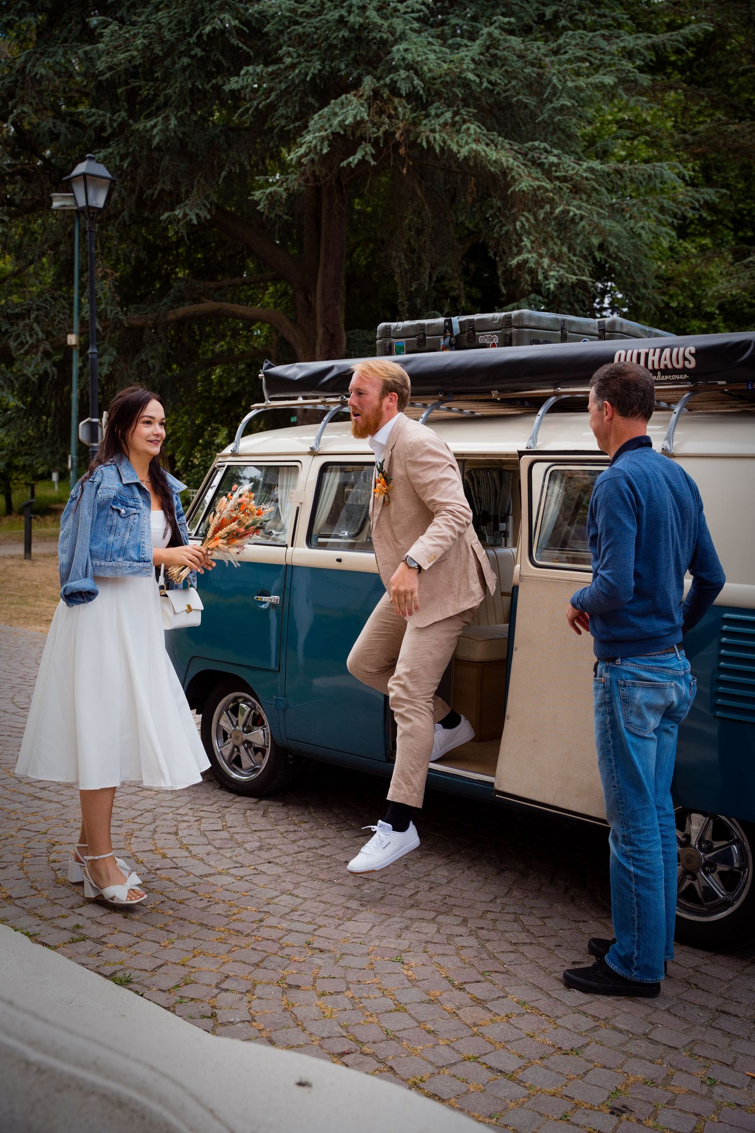 Bride and groom arrive at wedding venue in a vintage teal VW camper van in a tree-lined park.