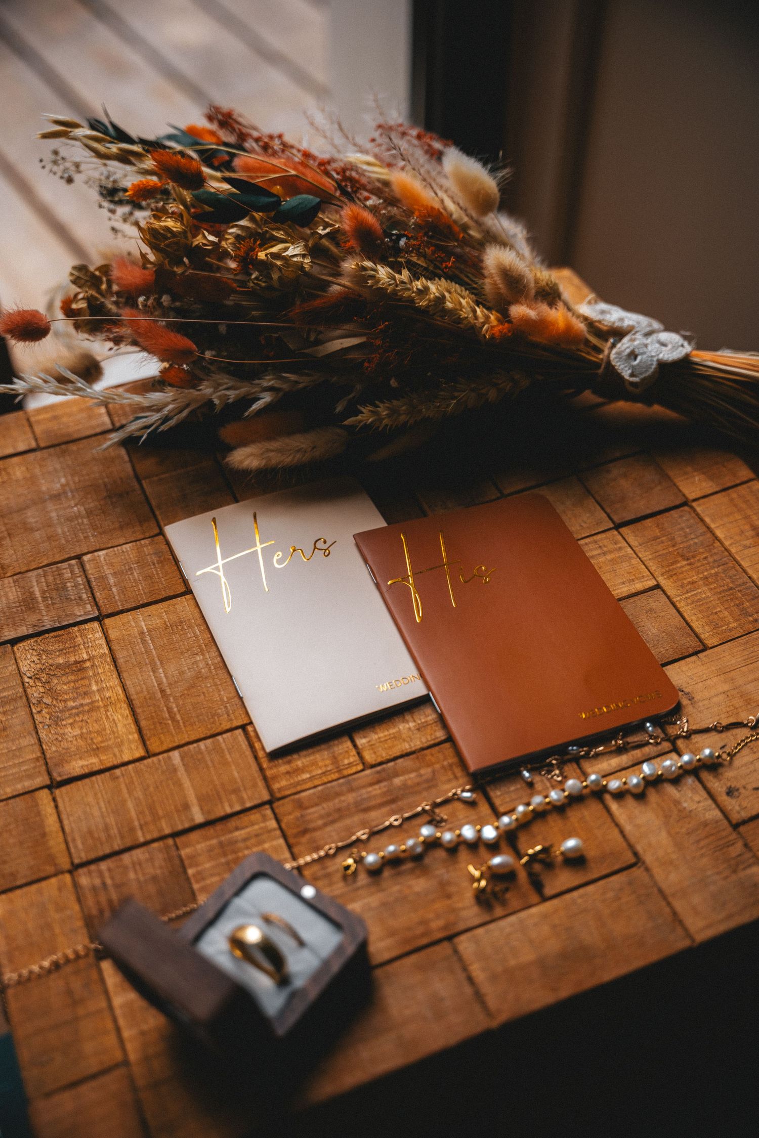 Wedding flat lay with dried floral bouquet, vow books, pearl bracelet, and ring box on wooden surface.