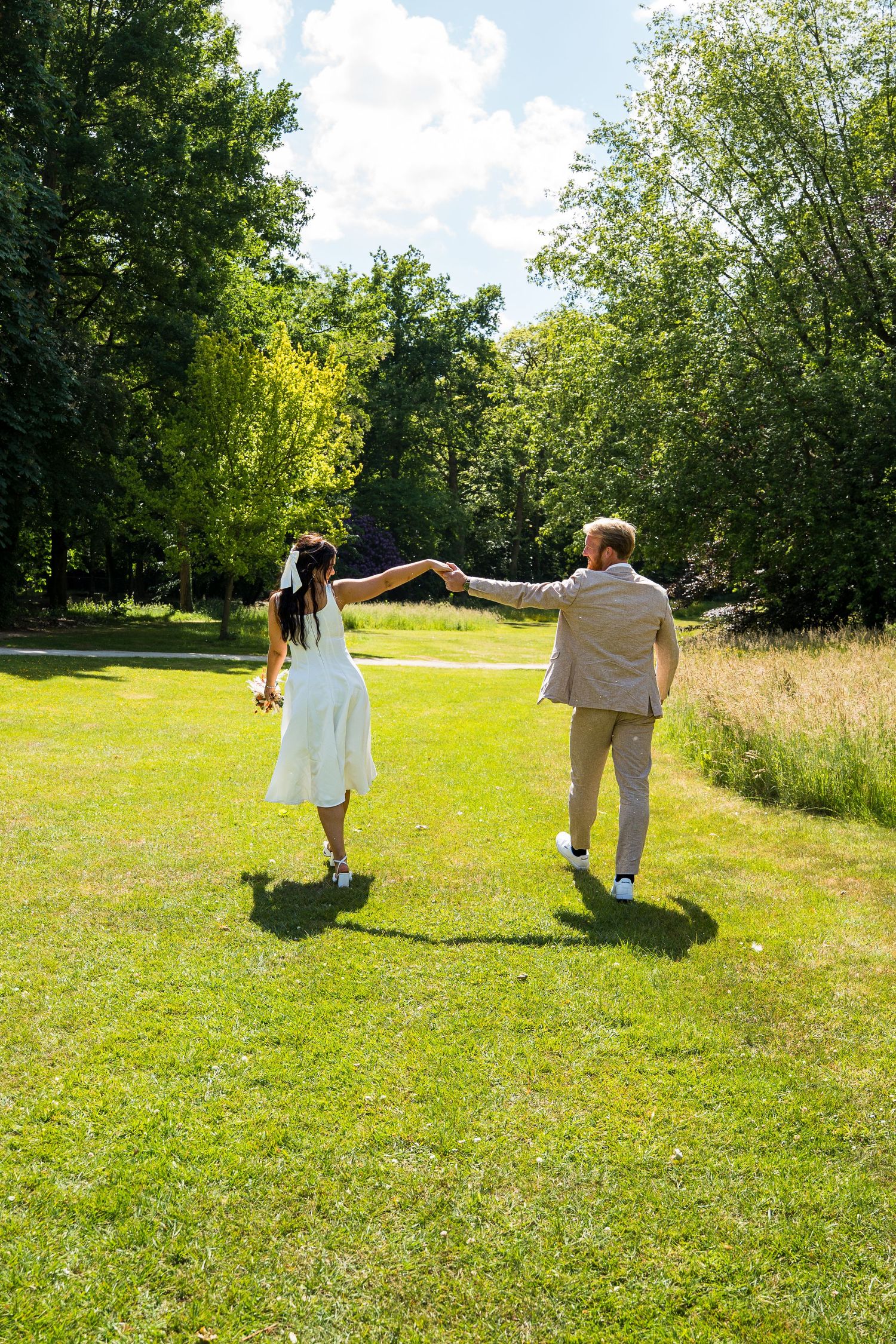 Bride and groom holding hands walking across a sunny green park on their wedding day.