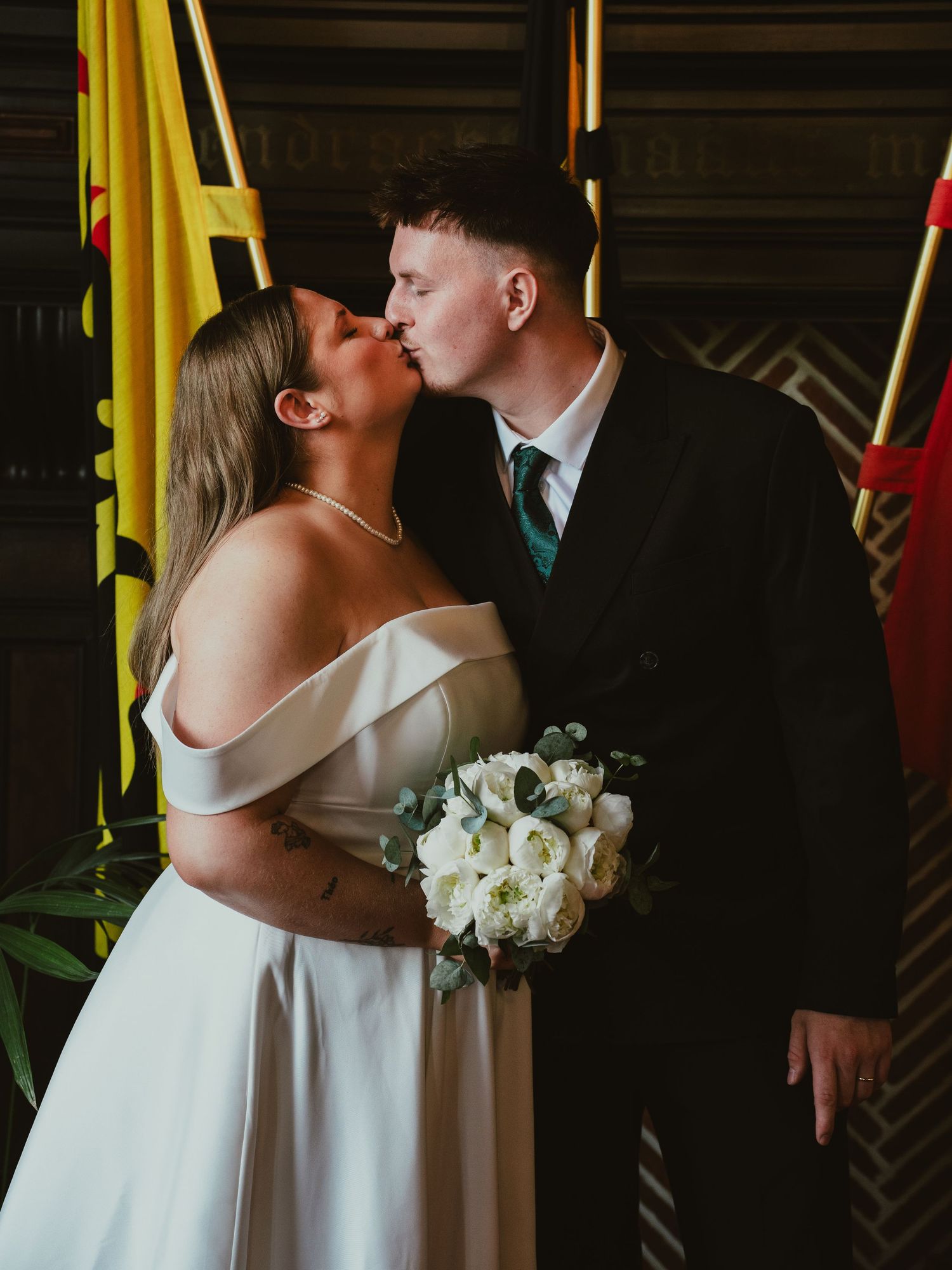 Newlyweds share first kiss at wedding ceremony with colorful flags in background.