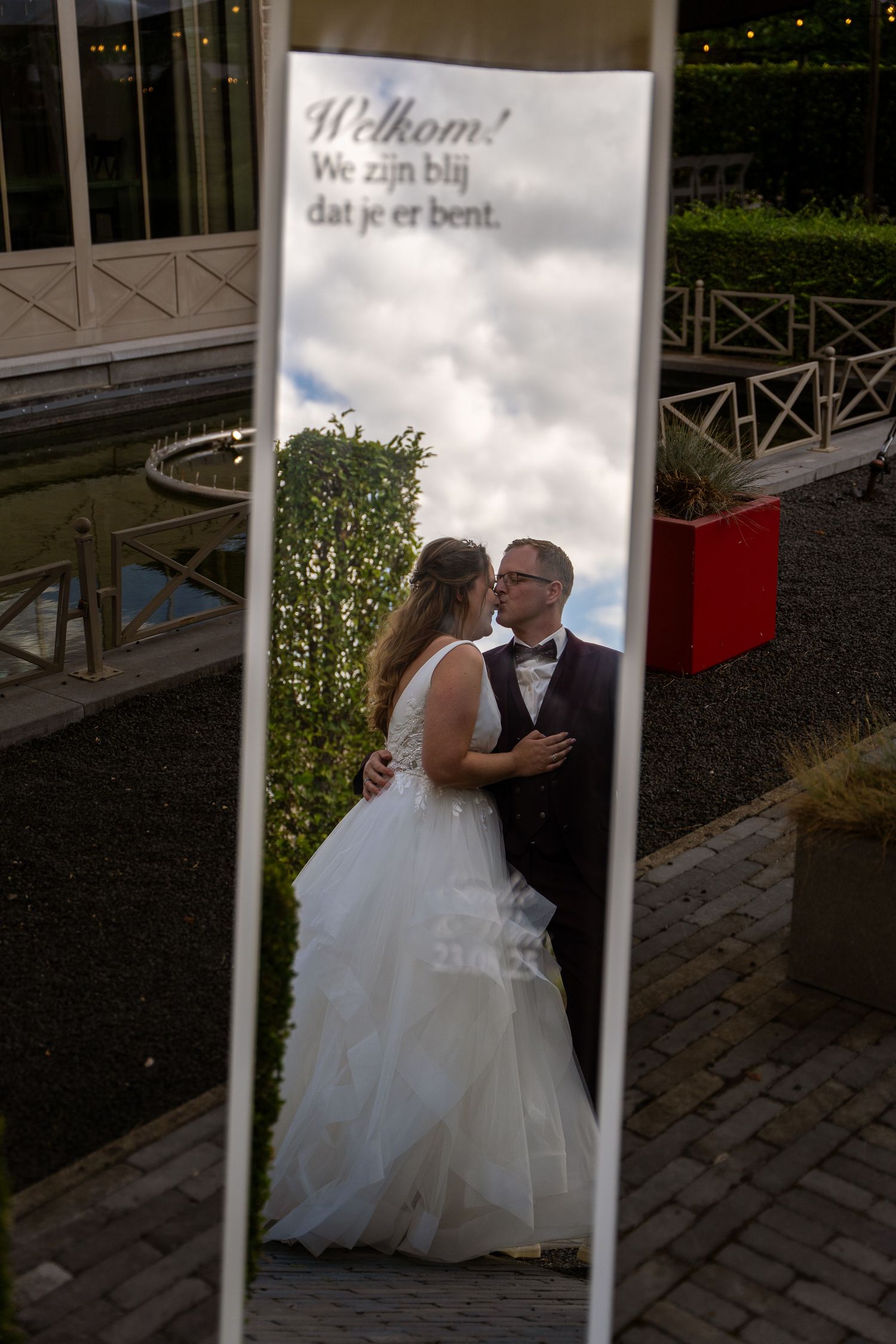 Bride and groom share a kiss reflected in a mirror displaying Dutch welcome message outdoors.