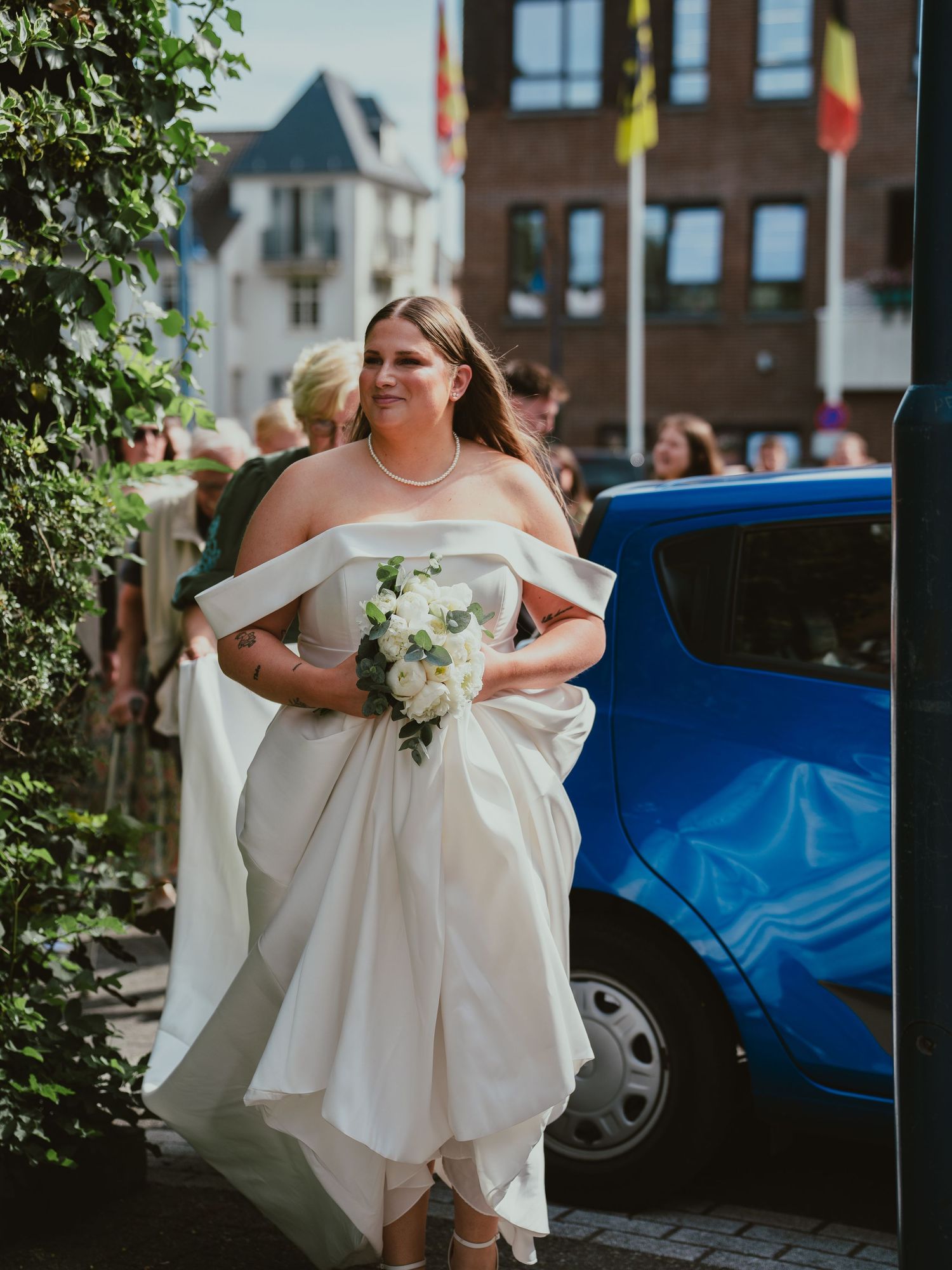Radiant bride in off-shoulder white gown carries bouquet arriving at wedding venue.