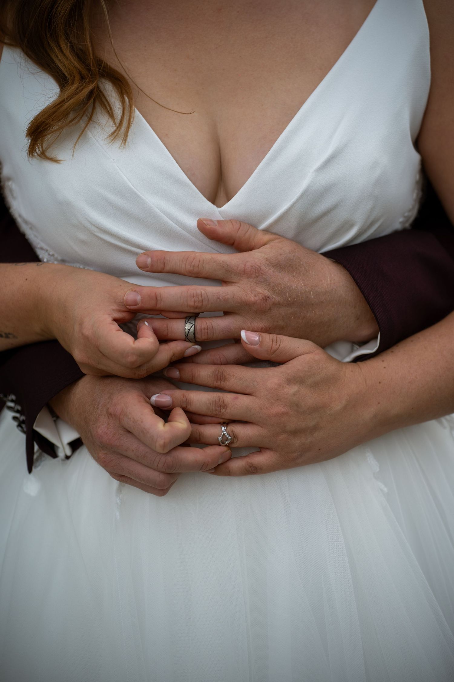 Close-up of bride and groom holding hands showing matching wedding rings over white bridal gown.