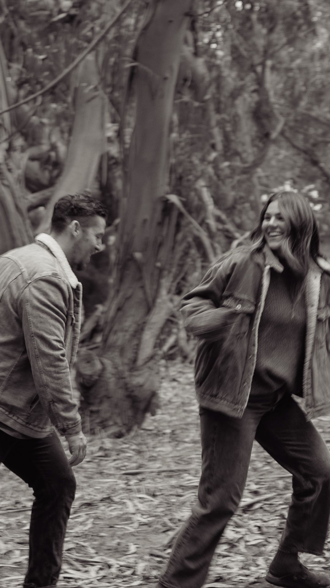 A couple laughs together in a black and white forest photo, both wearing casual jackets.