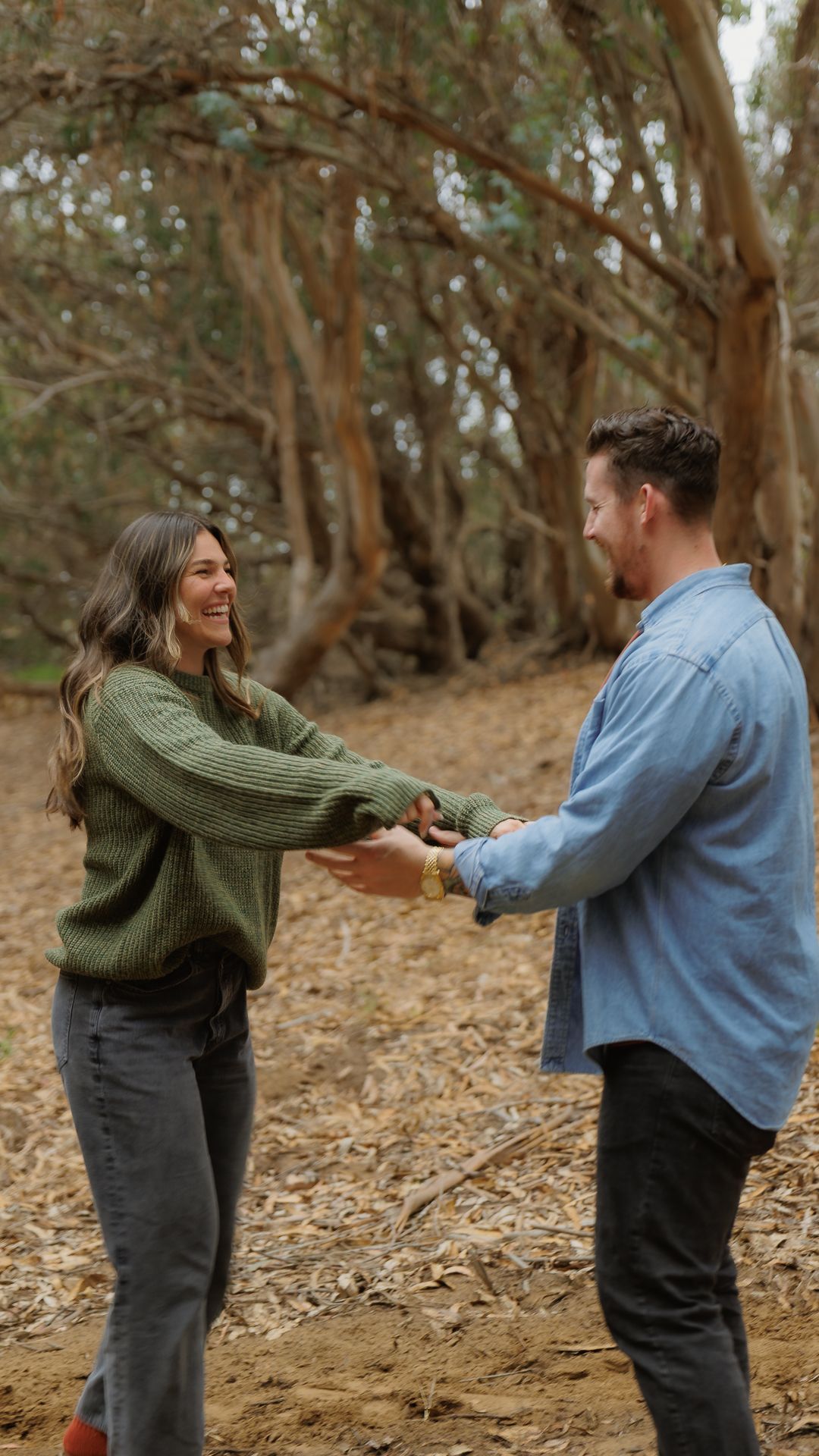 A smiling couple holds hands playfully on a leaf-covered forest path surrounded by trees.
