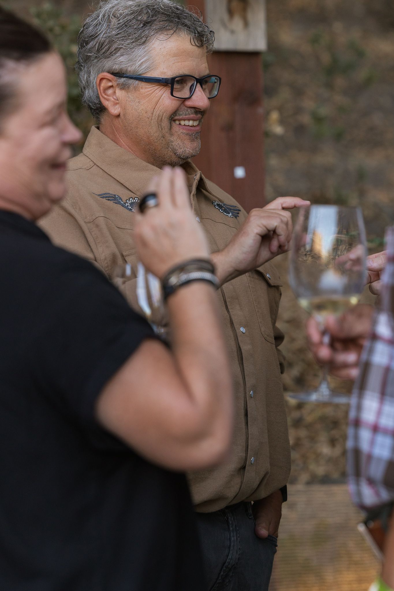 Man in glasses and brown shirt smiling at outdoor wine tasting event with guests holding white wine glasses.